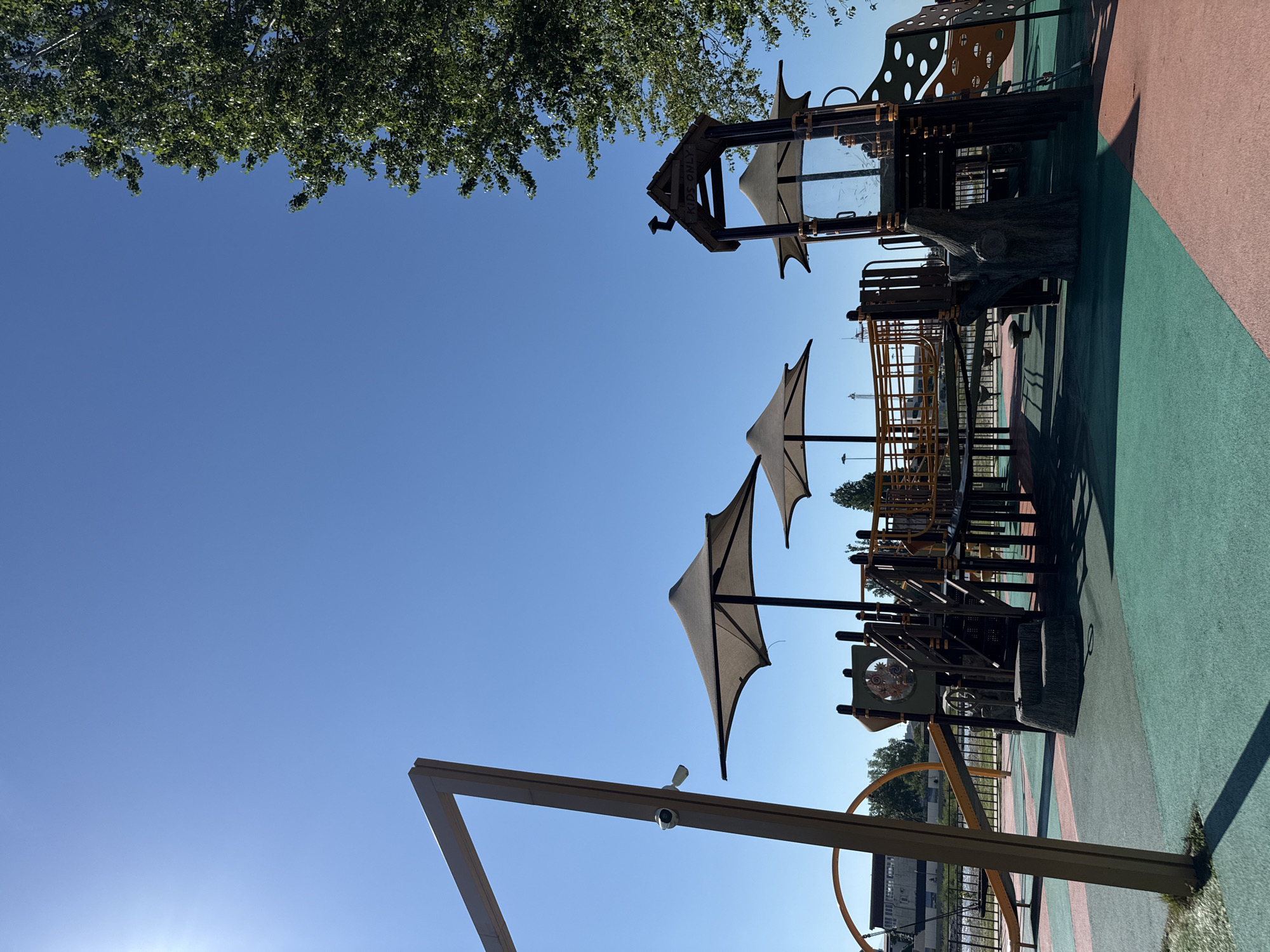 An outdoor playground is depicted under a clear blue sky with the sun shining brightly. The playground features a multi-level structure with slides, climbing elements, and shaded areas created by large, dark canopies. The ground surface is a mix of green and pink rubber. In the foreground, a large wooden frame with a security camera is visible. To the right, the branches of a large green tree extend into the frame. In the background, a body of water can be seen, with industrial buildings and structures on the opposite bank. The scene is devoid of people.
