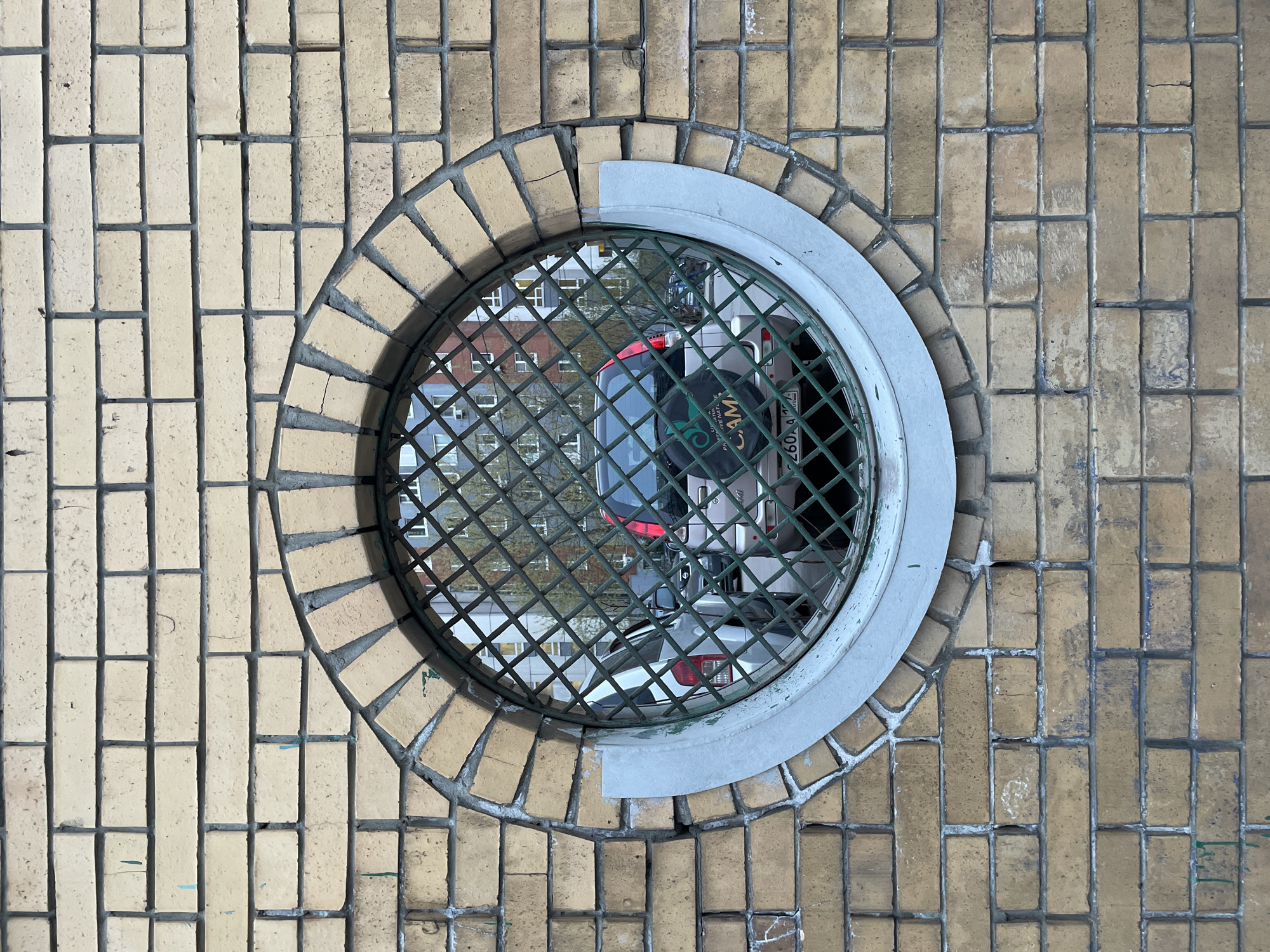 An outdoor scene shows a circular metal grate set into a tiled surface. The tiles are small, rectangular, and tan-colored with dark grout lines. The grate has a pale gray metal rim with two flat sections along the top edge, presumably for stability or reinforcement. Through the grate, a blurry reflection of urban elements is visible, including buildings, trees, and at least one vehicle. The reflection suggests the scene is likely a manhole cover or a similar feature embedded in a pedestrian area. License plate on the vehicle reads "2604". The image is rotated ninety degrees from the camera's perspective.