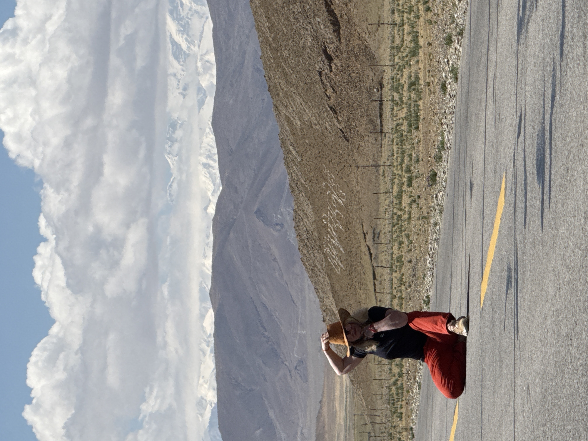 The image depicts a vast, open mountainous landscape under a bright, partly cloudy sky. In the foreground, a paved road with a single yellow dividing line on its right edge stretches into the distance. A person is standing on the road, slightly to the left. They are wearing a black top, loose red pants, and light-colored shoes. They hold a light-brown straw hat with both hands, partially obscuring their face as they look forward towards the mountains. Long, distinct shadows are cast from the person and other unseen objects onto the road, indicating bright sunlight, likely during the late morning or afternoon.
The terrain immediately adjacent to the road is dry and arid, covered with sparse, light-brown vegetation and small rocks. Faint utility poles or fence posts are visible along the roadside to the right.
The background is dominated by majestic mountains. A closer range appears rocky and barren, in shades of grey and brown. Beyond these, towering snow-capped peaks stand prominently against the clear blue sky, partially adorned with large, fluffy white clouds, particularly on the left side.
The scene conveys a sense of grandeur and isolation, characteristic of high-altitude mountain regions. The provided context suggests the location is Mainpu township, China, which aligns with the depicted landscape. There is no visible text in the image.