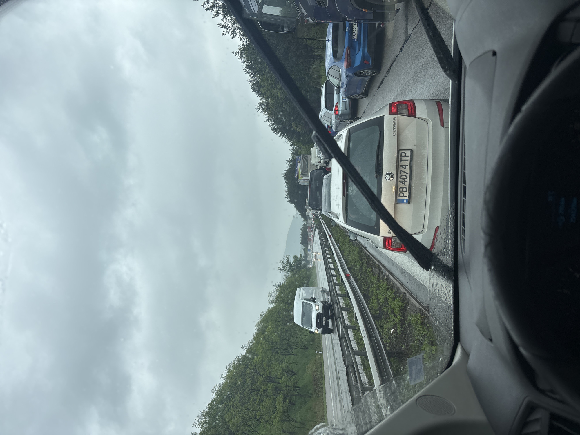 The image shows a traffic jam on a highway near Gorna Vasilitsa, Bulgaria. It is a cloudy day. Cars are lined up bumper to bumper in the right lane of the highway. A white Skoda Octavia with license plate PB 4074 TP is visible in the foreground. A white van is traveling in the left lane. The view is from inside a vehicle, looking through the windshield. Raindrops are visible on the windshield. Trees line the side of the highway.