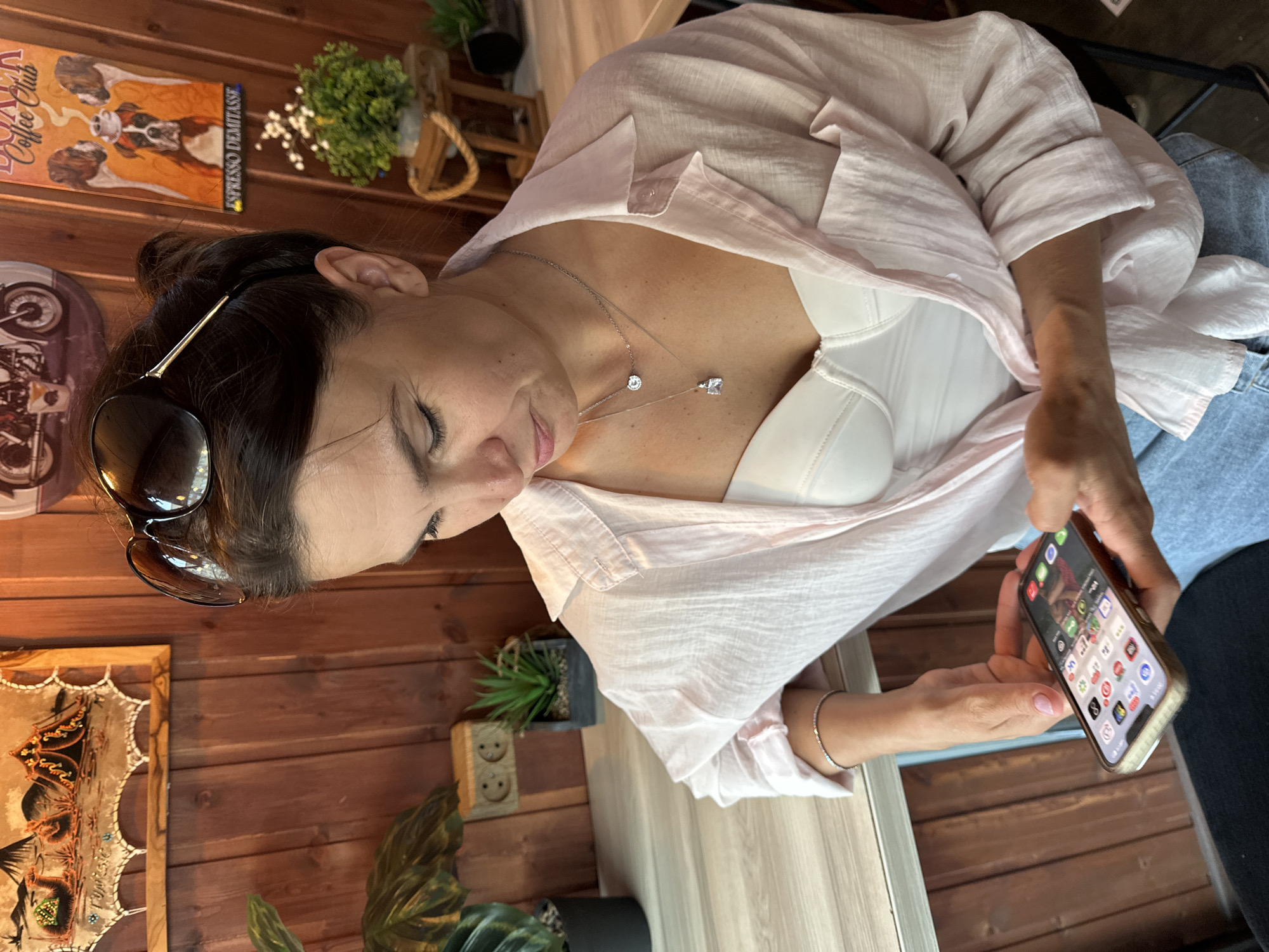 A woman is sitting at a table, looking down at her phone. She is wearing a light pink unbuttoned shirt over a white bustier and blue jeans. She has sunglasses on top of her head and is wearing two necklaces. The background is a wooden wall decorated with various posters and pictures, and there are plants on shelves. The scene appears to be indoors, possibly a cafe or restaurant. The image is taken in Petropavlovsk-Kamchatsky, Russia.