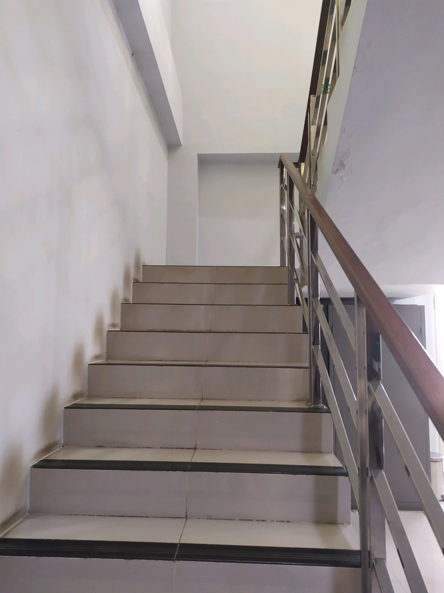 A concrete staircase with gray tiled steps and black nosing ascends upwards to the right, bordered by a metal railing with a wooden handrail. The walls are painted white, and the ceiling is also white. The environment appears to be an indoor common area, possibly in a building in Wates, Indonesia, though no specific identifying features of Wates are visible. The scene is static with no people or ongoing actions.