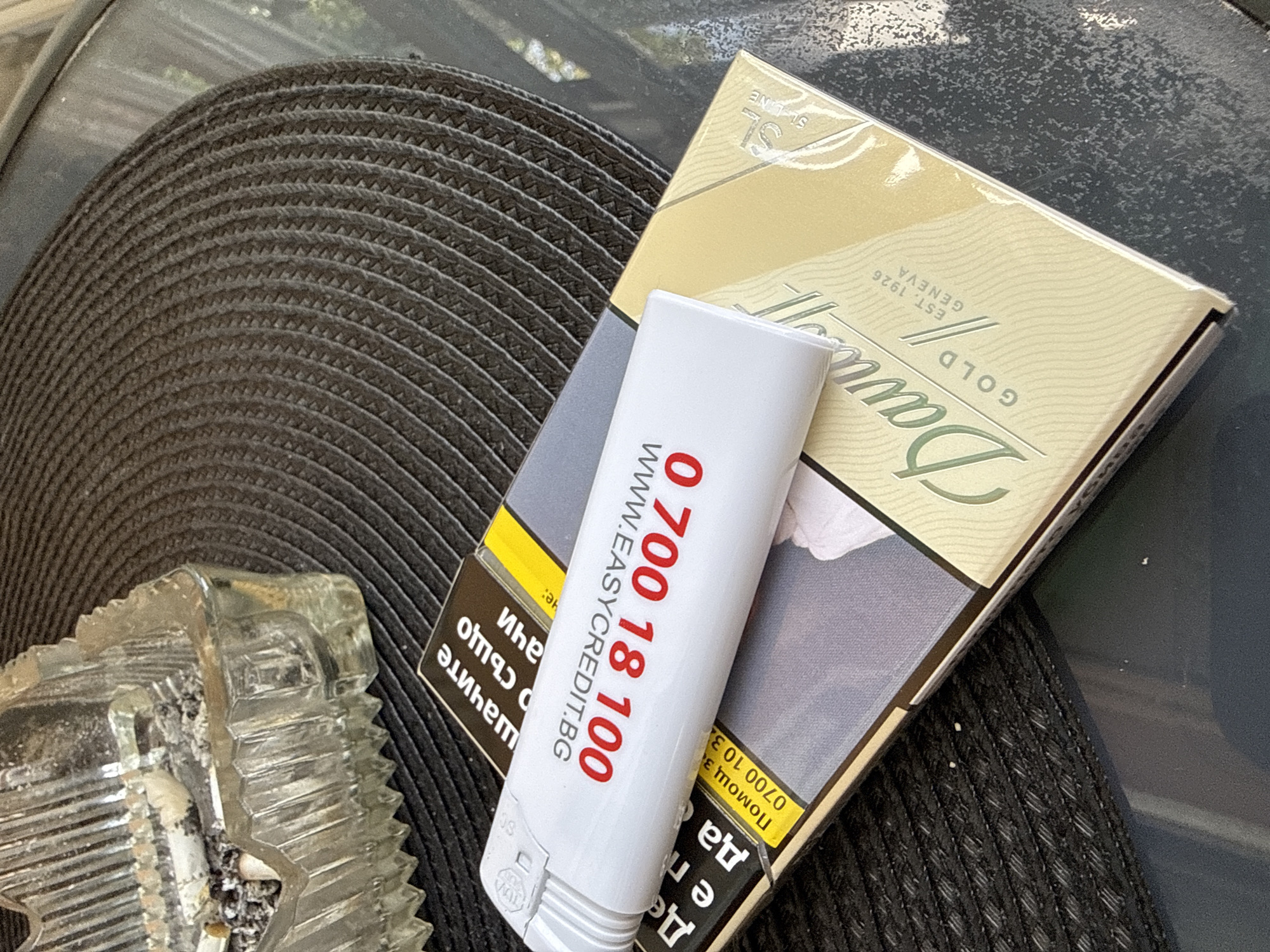 The image shows a pack of Davidoff Gold cigarettes and a white lighter with red lettering on a dark woven surface, possibly a car dashboard. An ashtray containing cigarette butts is visible to the left. The lighter displays the phone number "0 700 18 100" and the website "WWW.EASYCREDIT.BG." The cigarette pack is gold and white, with the brand name "Davidoff" and the text "GOLD GENEVA EST. 1926." The overall scene suggests smoking. Given the website displayed on the lighter, the location might be Lovech, Bulgaria, where Easy Credit operates.