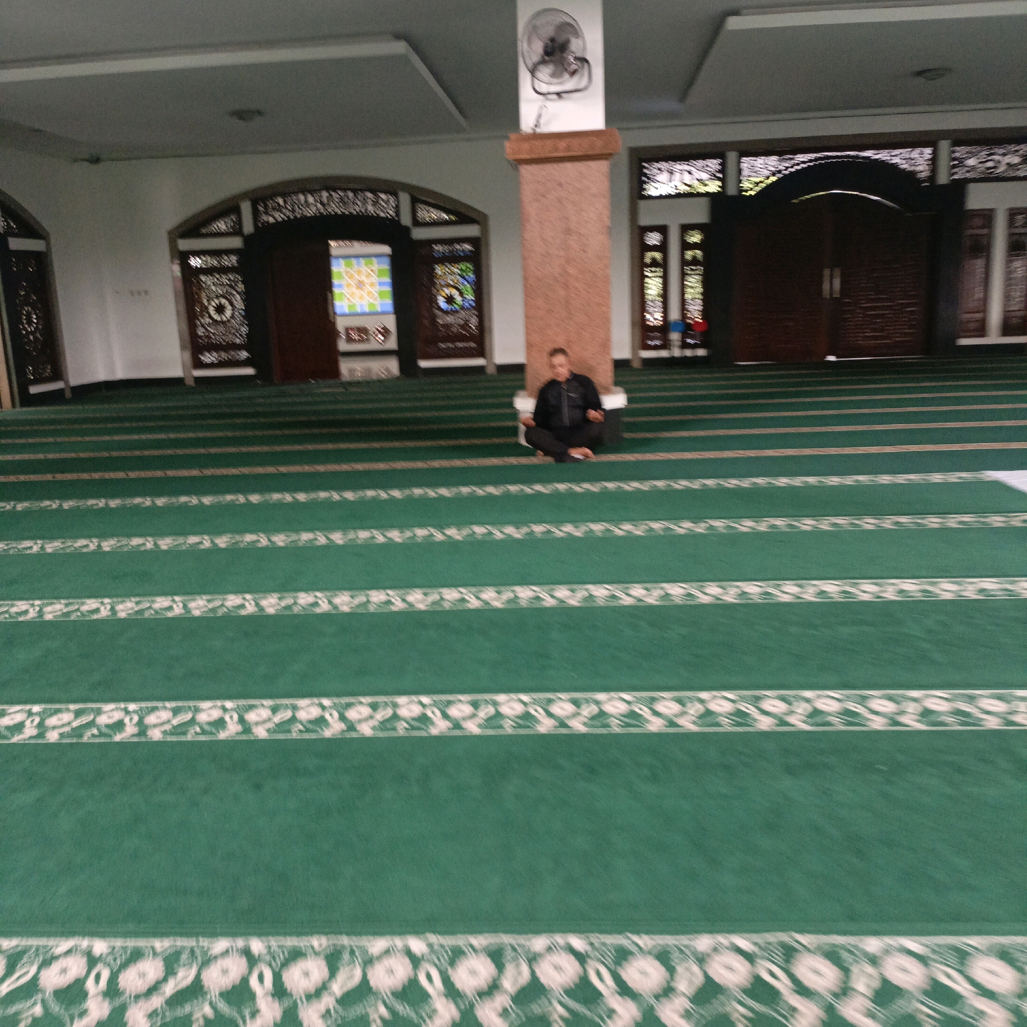 The image shows the interior of a mosque with green carpets laid out in rows. A person is sitting cross-legged on the carpet, leaning against a pillar. The mosque features arched doorways with decorative elements and stained glass windows. A ceiling fan is visible on the pillar. The overall environment suggests a place of worship.