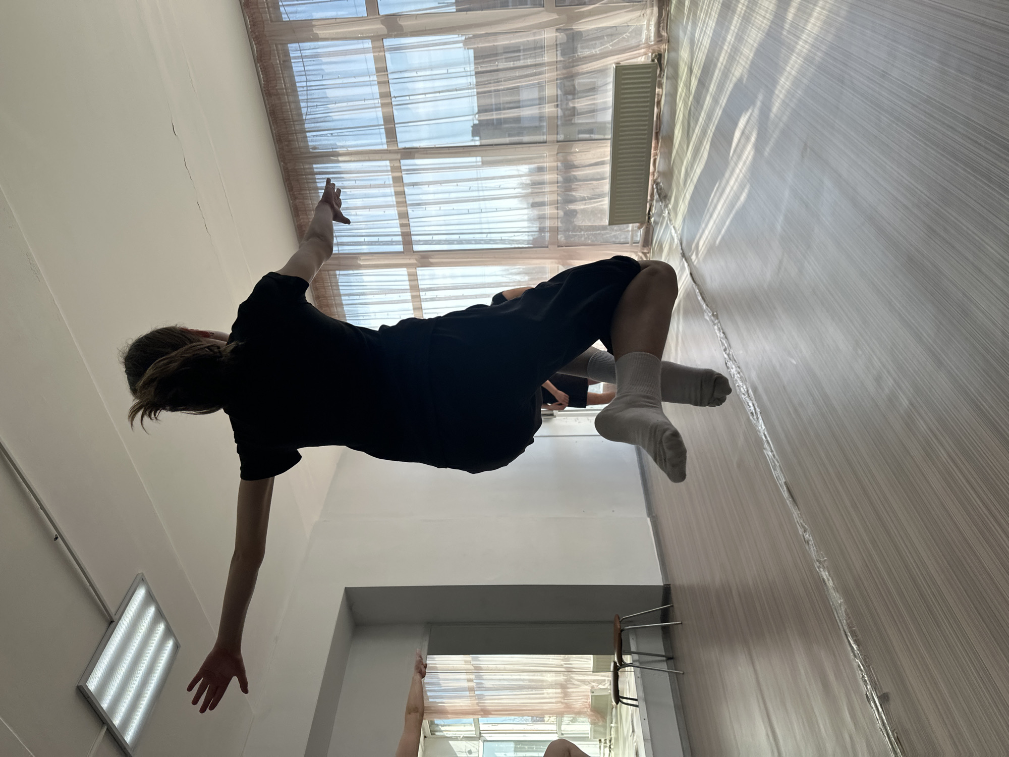 A person is seen from a low angle, positioned upside down relative to the viewer's perspective. The person wears a black t-shirt and dark shorts, with white socks on their feet. Their right arm is extended upwards, while the left arm hangs downward.
The background includes a room interior with a window covered by light-colored curtains, suggesting daylight. A heating radiator is visible near the window. The walls appear to be a light color. The floor has a wood-like pattern. In the lower part of the image, another person's arm is partially visible near a doorway or opening.
Without additional information, it is not possible to confirm the precise location in Votkinsk, Russia.