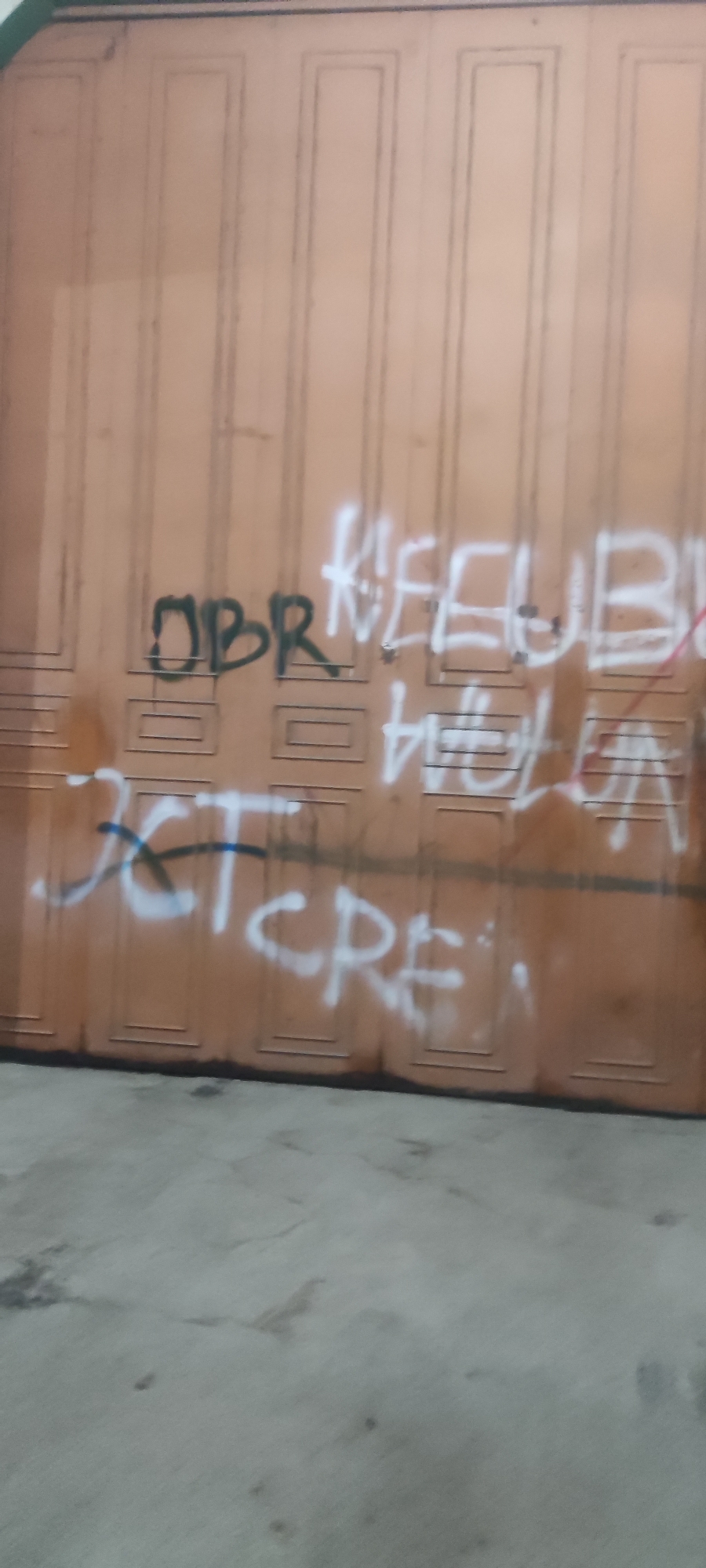 The image depicts a close-up of a weathered, light brown garage door with vertical paneling. The door is covered in graffiti. On the left side, the word "OBR" is spray-painted in dark green. To the right of "OBR," white spray paint forms the words "KEEUBA" and "HELA" which are partially obscured by other spray paint marks. Further to the right and lower down, a blue line is visible intersecting with white spray paint that spells out "JCTCRE". The lower portion of the image shows a grey concrete ground surface. The scene appears to be outdoors, possibly in an alley or street within Gebang, Indonesia, as indicated by the context provided.
