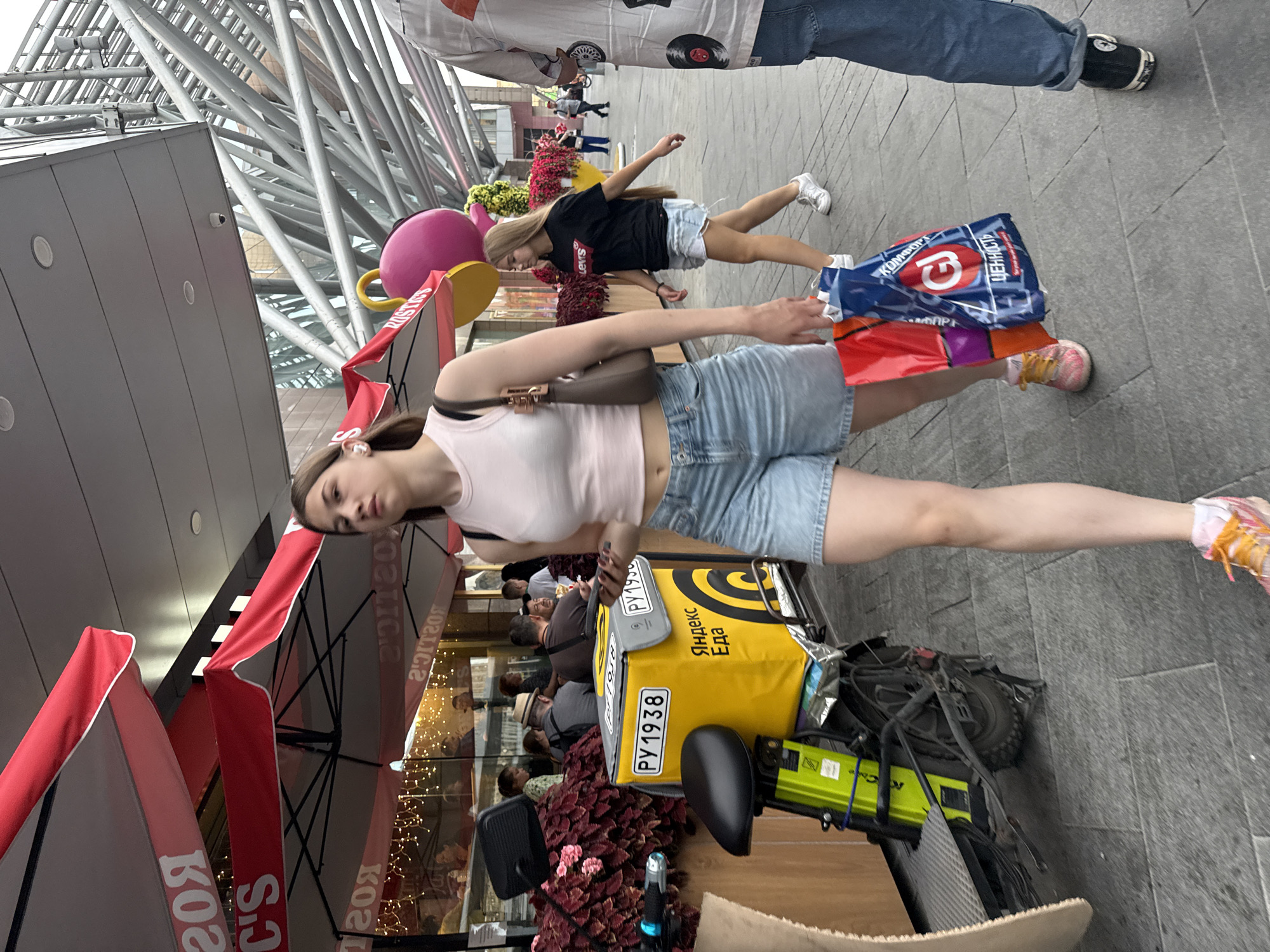 The image captures a street scene in Moscow, Russia, likely a commercial area with outdoor dining and shops. In the foreground, a young woman with blonde hair is walking towards the camera, wearing a light pink tank top and denim shorts. She carries a brown shoulder bag and two shopping bags, one blue with red and white text and graphics, and another red bag partially visible. To her left, a bright yellow delivery box with Russian text "Яндекс Еда" (Yandex Food) is attached to a green electric scooter, parked on the sidewalk. In the middle ground, another young woman with long blonde hair is walking away from the camera, wearing a black t-shirt with a white "Levi's" logo and light denim shorts. Behind her, several people are seated at tables outside a building, with red awnings overhead featuring white lettering that appears to say "ROSTICS". The background shows modern architecture with a metallic, skeletal structure and glass facades. The ground is paved with gray interlocking stones. The overall atmosphere suggests a bustling urban environment.