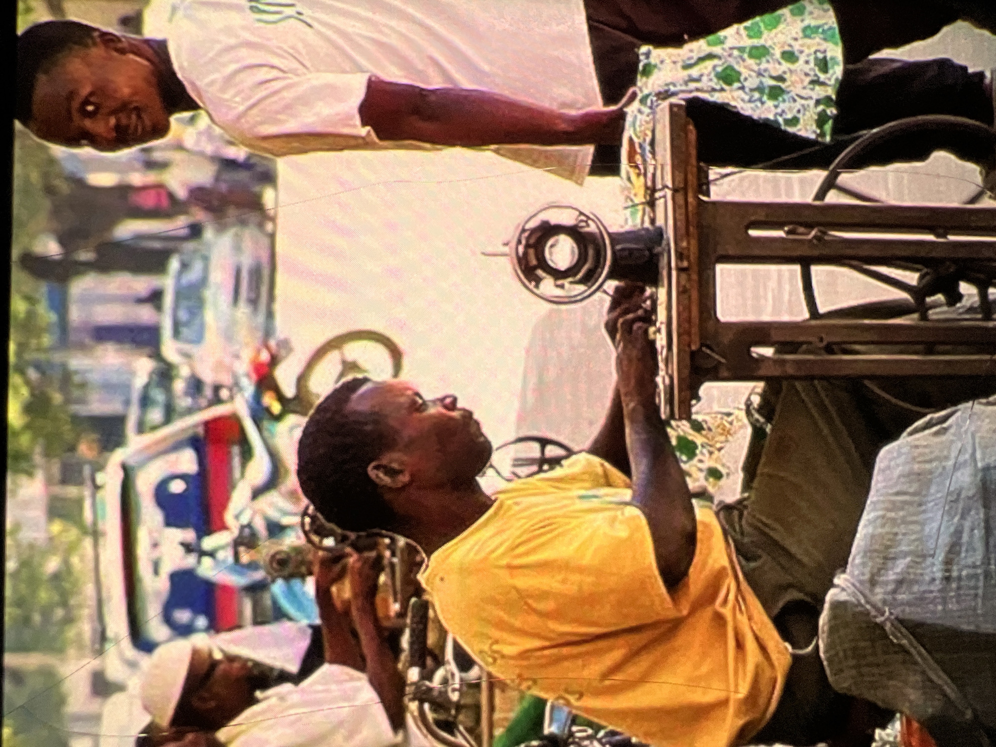 The image depicts an outdoor scene in Lagos, Nigeria, likely a busy street or market area, with a focus on a tailor working at a sewing machine. In the foreground, a man wearing a bright yellow t-shirt and dark trousers is seated and actively engaged with a vintage sewing machine. His hands are positioned on the machine, suggesting he is in the process of sewing. The sewing machine itself is mounted on a wooden table with a wheel attached to its side. A patterned fabric, predominantly green and white, is draped over the table and partially covers the sewing machine.
To the right of the tailor, another individual, a young man, is partially visible, looking towards the camera with a slight smile. He is wearing a white t-shirt. Behind them, the background shows a blurry street scene with vehicles, including a red and blue car, and other people. The overall atmosphere suggests a vibrant, active urban environment. The lighting indicates it is daytime.