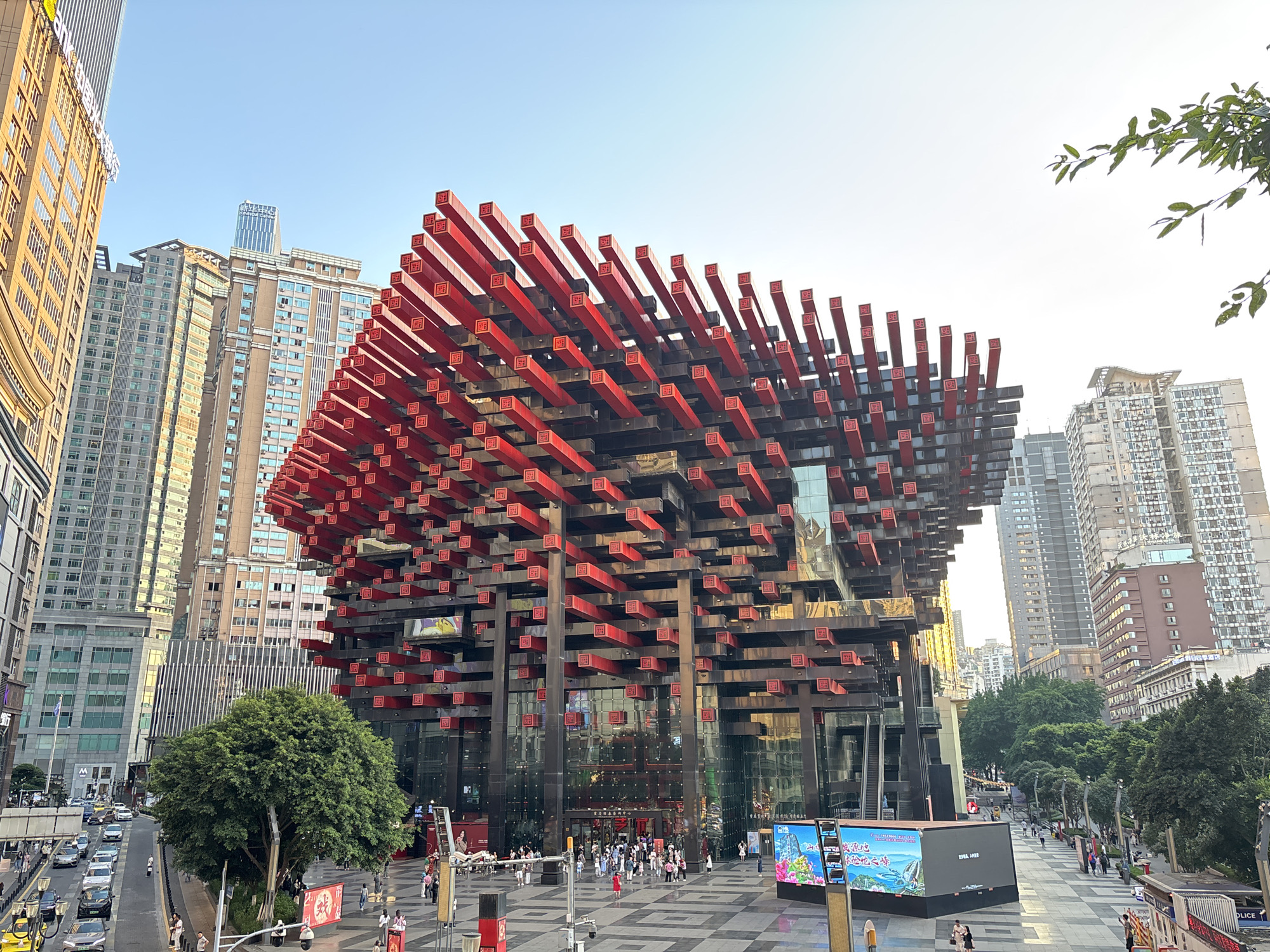 The image shows the Raffles City Chongqing complex, located in the Yuzhong District of Chongqing, China. The foreground features a plaza with pedestrians, trees, and street furniture. The Raffles City complex stands prominently with its distinctive design of horizontal red and black structural elements. The facade of the building appears to be primarily glass. Other buildings are visible in the background. Vehicles travel along a street on the left. The sky is clear.