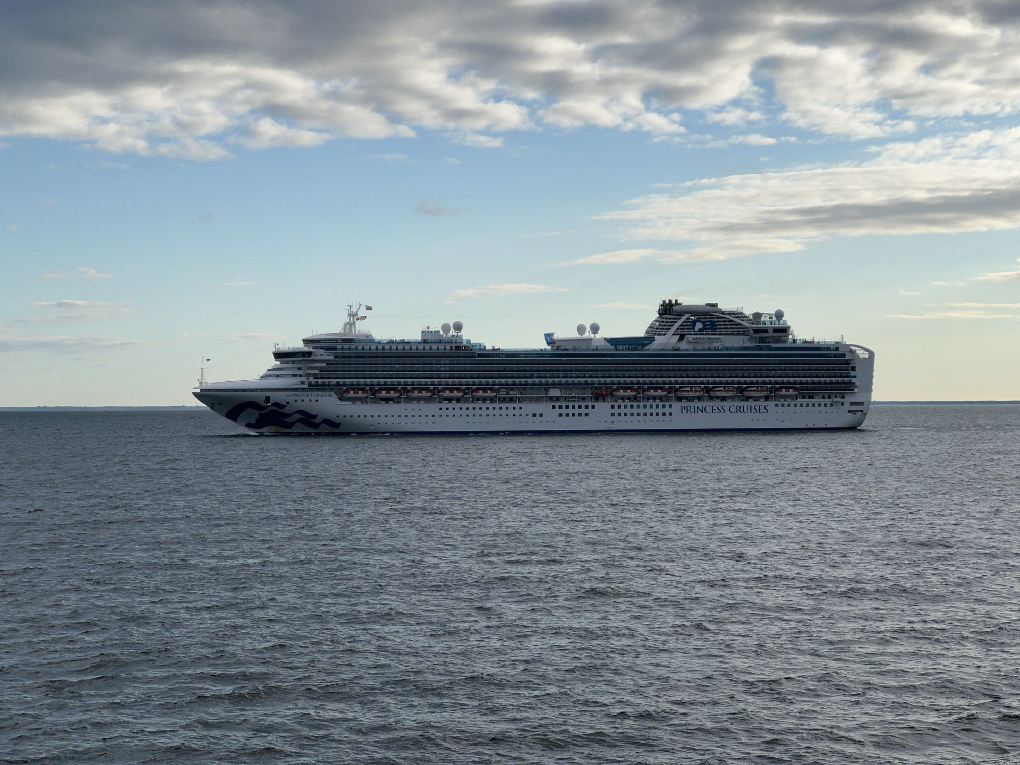 A large white cruise ship, the "Sapphire Princess" of Princess Cruises, sails on a calm, dark blue sea under a partly cloudy sky. The ship is positioned in the center of the frame, moving from left to right. It is a multi-deck vessel with numerous windows visible along its sides, and life rafts are stowed on the upper decks. The name "PRINCESS CRUISES" is emblazoned on the side of the ship. In the distance, a faint landmass is visible on the horizon. The scene is one of maritime transit, with no people or specific city landmarks clearly identifiable in the immediate vicinity of the ship or the water. The presence of the "Sapphire Princess" in Polish waters suggests it might be in the Baltic Sea, possibly near the port of Gdynia, Poland, given its capacity for large vessels.
