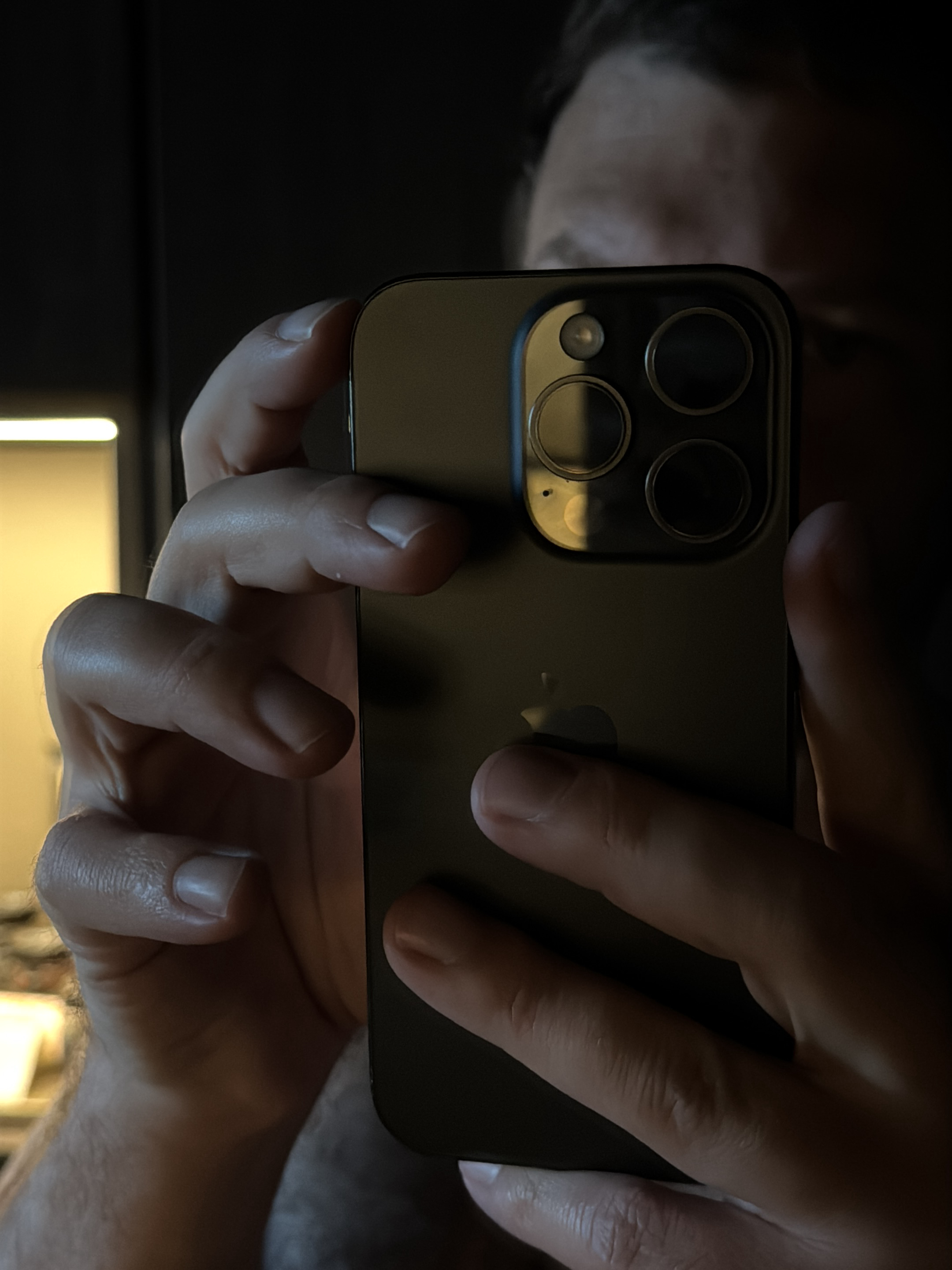 The image is a close-up shot of a person holding a dark-colored smartphone. The phone's camera module, featuring multiple lenses, is visible and reflects light, suggesting it might be an iPhone. The person's hands are wrapped around the phone, holding it in a portrait orientation, implying they are either taking a photo or using the device. The background is dark and out of focus, with a blurred bright light source on the left side, possibly an artificial light or a window. The overall lighting is low-key, with harsh shadows. There is no discernible information in the image that places the scene in Milan, Italy.