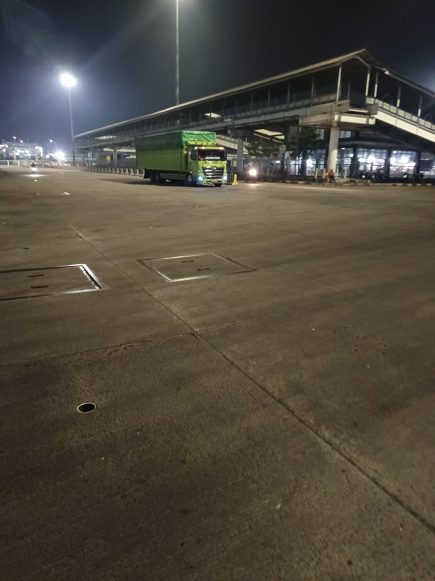 A green truck is parked on a paved lot at night in Bakauheni, Indonesia. The truck is facing right and is mostly visible, with its trailer covered by a tarp. In the background, there is a large, elevated pedestrian walkway or bridge structure with lights. Several streetlights illuminate the scene. The ground is concrete and appears to be a public area, possibly a terminal or transportation hub, with manhole covers visible. There are no people clearly depicted interacting with the truck or the immediate surroundings. The atmosphere is dark and illuminated by artificial lighting.