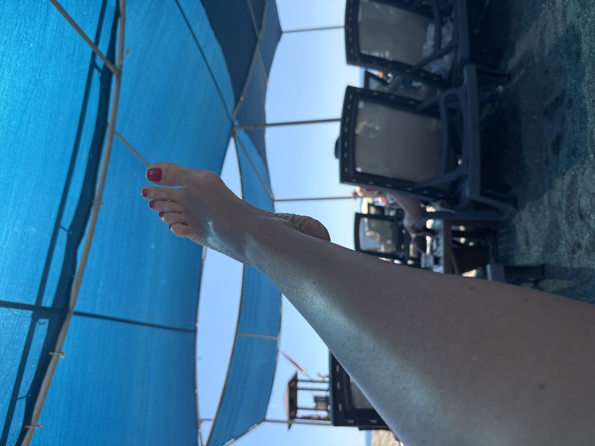 The image shows a scene at a beach in Unknown, Unknown. A person's leg and foot, with red nail polish on the toes, are prominently displayed in the foreground. Above, a blue beach umbrella provides shade. In the background, beach chairs are arranged on the sand. The sky is visible through the umbrella's gaps.