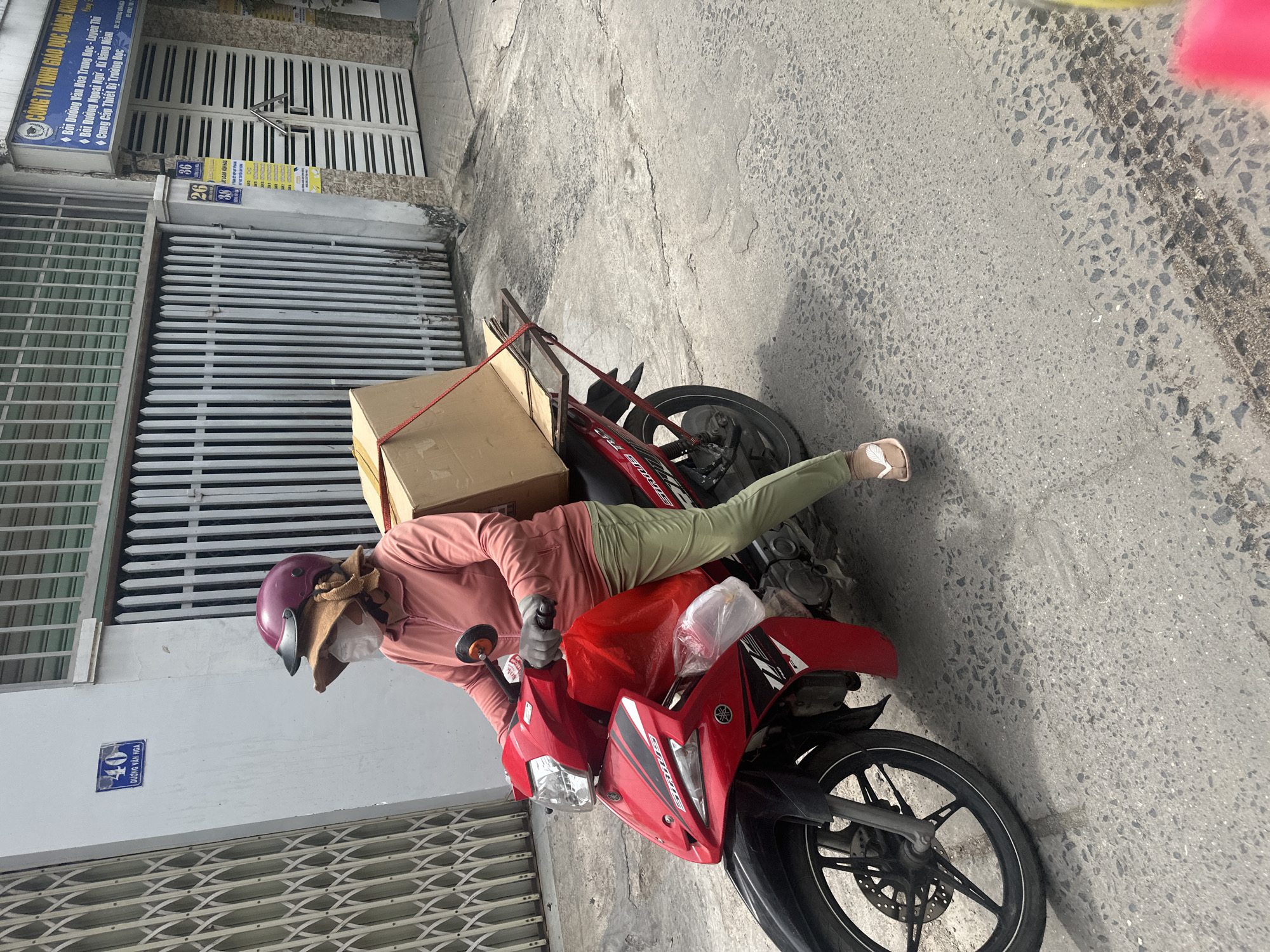 A person wearing a pink long-sleeved shirt, light green pants, a dark pink helmet, and a light-colored face mask is seen sitting on a red motorcycle parked on a paved street. The motorcycle appears to be carrying cardboard boxes secured to its rear. The location is in Bac Nha Trang Ward, Vietnam, as indicated by the Vietnamese text on nearby buildings at addresses numbered 26, 36, and 40 on Duong Van Nga street. The background includes buildings with closed metal gates and concrete walls.