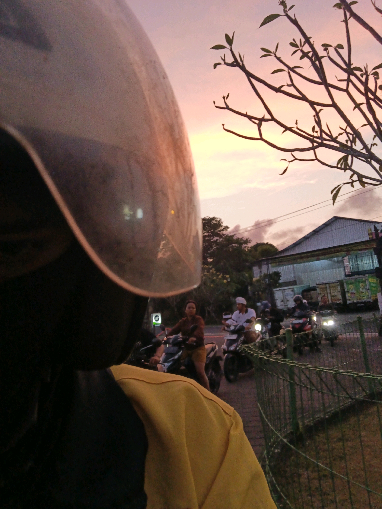 This is a first-person perspective, medium-shot photograph taken at dusk on a street in Denpasar, Indonesia. The foreground is dominated by the reflective surface of a motorcycle helmet, with the rider's yellow jacket visible on the right. In the midground, several people on motorcycles are moving along a paved road. One person in the center is wearing a white head covering. A building with a metal roof is visible in the background, along with a tree with sparse leaves silhouetted against the twilight sky. A green metal fence runs along the right side of the image, separating the road from a grassy area. The sky displays a gradient of pink and orange hues.