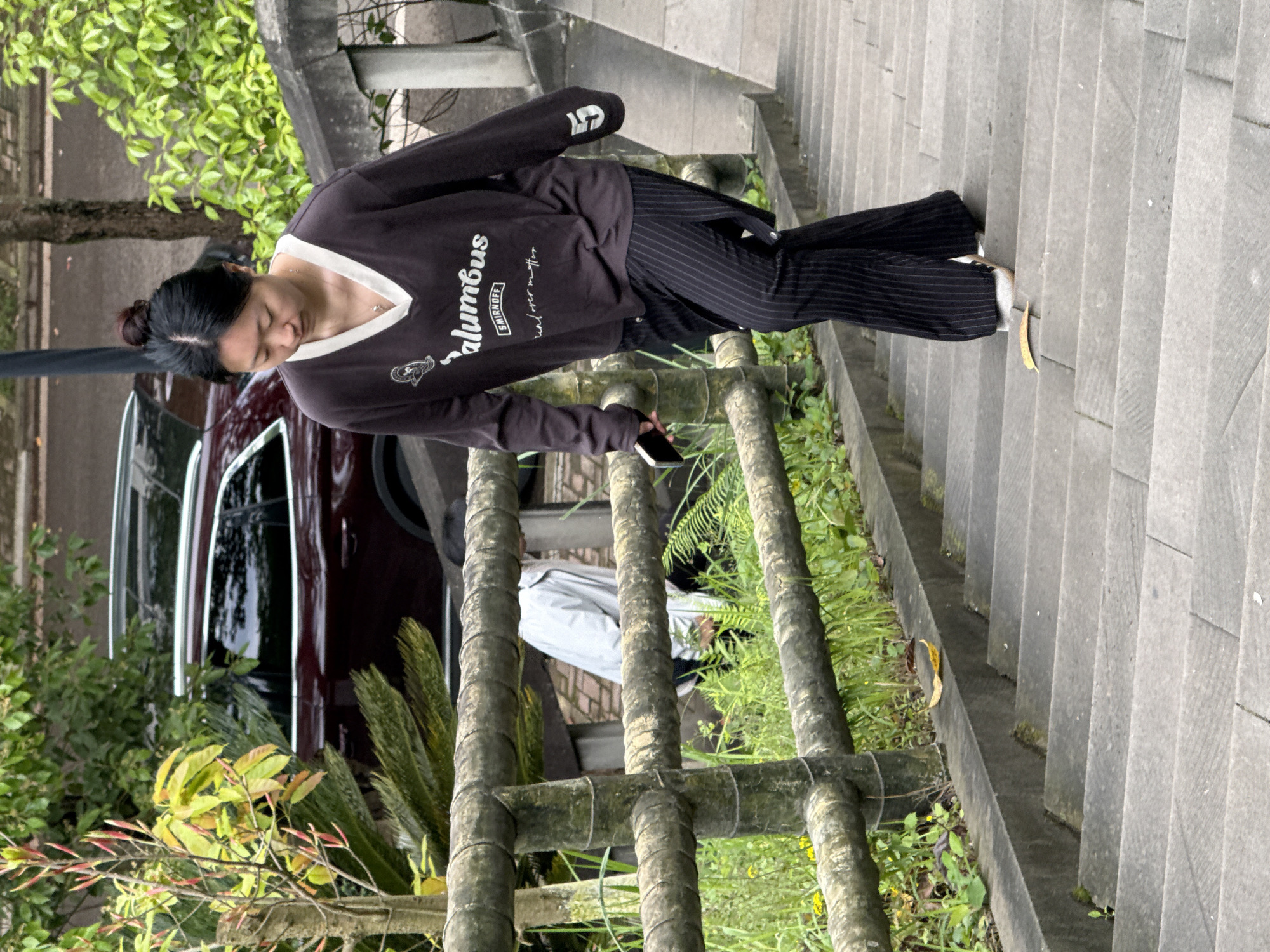 A person, likely female, stands on the right of stone steps outdoors. She is wearing a dark-colored, long-sleeved shirt or sweatshirt with a white v-neck and the word "Columbus" printed on the front. The person also wears vertically striped pants. She appears to be holding a smartphone in her hand.
The steps are constructed of gray stone and appear to be part of a staircase. To the left of the person is a weathered, wooden railing and green foliage. A maroon vehicle and greenery are visible in the background. The scene is likely in Jiangjin, China.