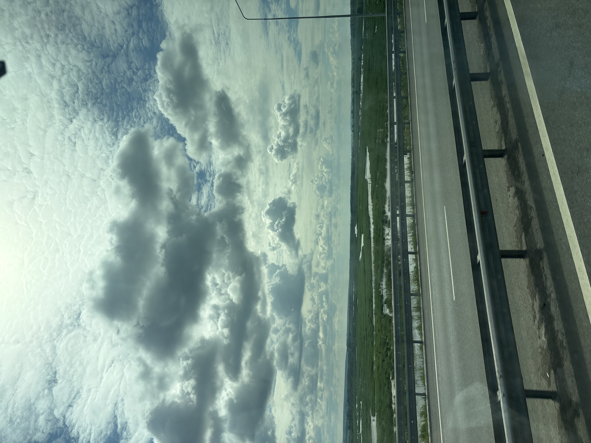 The image captures a view from a moving vehicle on a highway, likely in the vicinity of Menzelinsk, Russia, given the landscape. A two-lane highway with a metal guardrail dominates the foreground. Beyond the highway, a vast expanse of green marshland dotted with bodies of water stretches into the distance. The sky above is filled with dramatic, gray and white clouds, suggesting a dynamic weather pattern. A single lamppost is visible on the right side of the image. The overall scene conveys a sense of travel through a rural or semi-rural landscape.