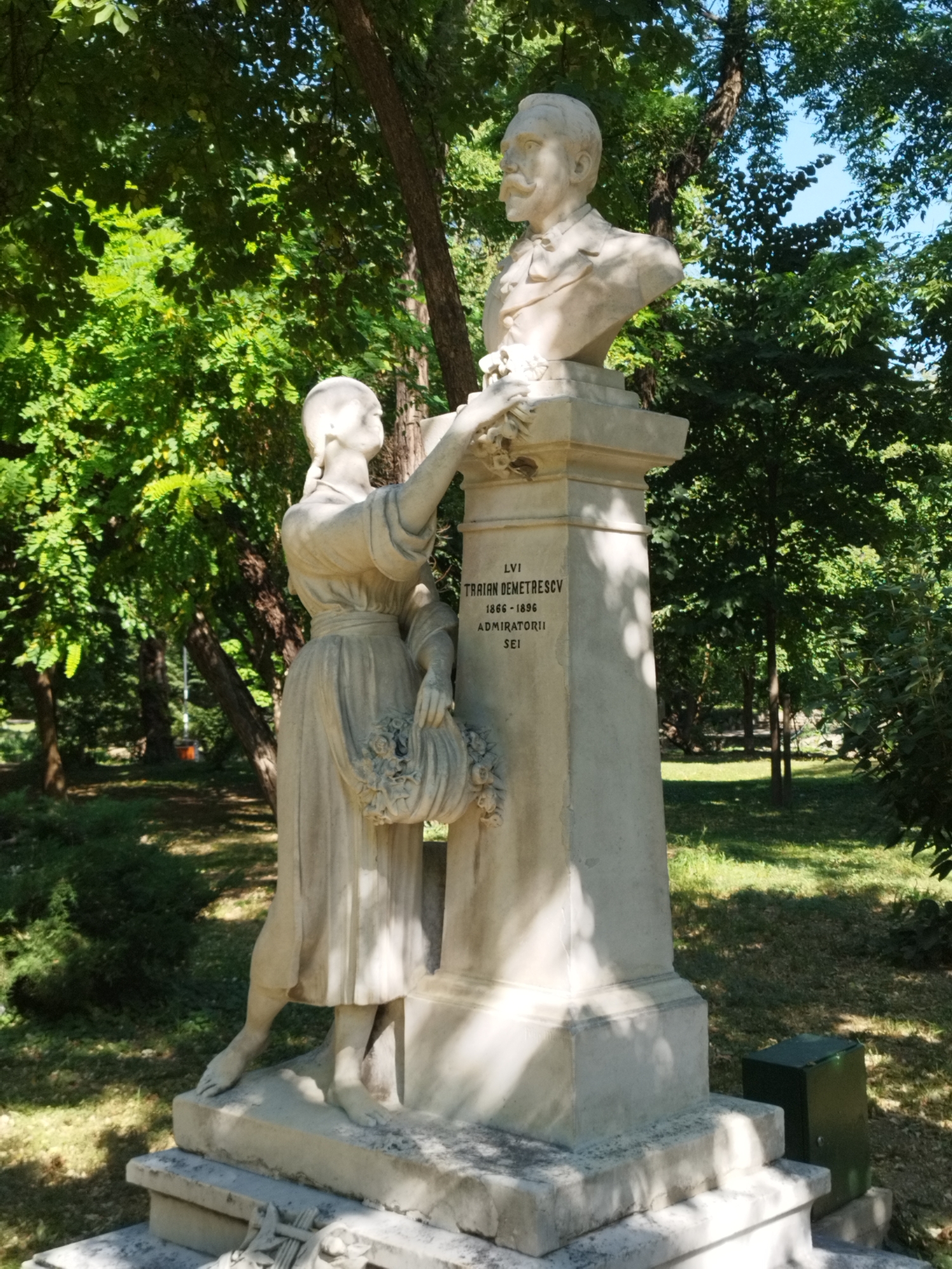 The image depicts a white stone monument featuring a bust of a man with a mustache, wearing a suit and a bow tie. Beside the bust, a female statue in a draped dress holds a basket of flowers. The monument has an inscription that reads "LVI TRAIAN DEMETRESCV 1866-1896 ADMIRATORII SEI". The monument is situated outdoors in a park-like setting with abundant green trees providing shade. The ground is covered with grass and scattered leaves. In the background, more trees and a glimpse of blue sky are visible. A dark green utility box is positioned to the right of the monument's base. This monument is located in Bucharest, Romania, specifically within the Herăstrău Park (now King Michael I Park), and honors Traian Demetrescu, a Romanian poet and critic.