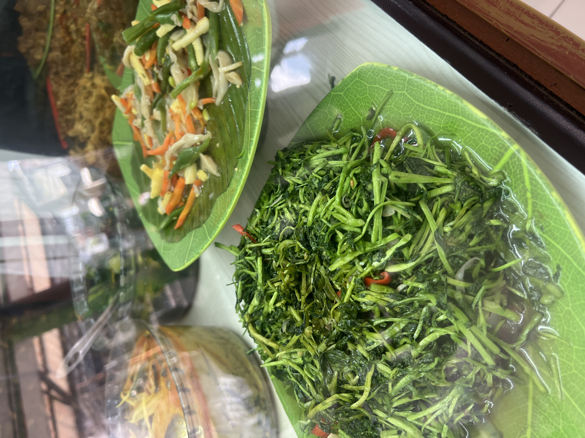 Two plates of Indonesian cuisine are displayed behind glass, likely in a food stall or restaurant in Kuta, Indonesia. The plate in the foreground is filled with a pile of sautéed green vegetables, possibly water spinach or kangkung, with visible red chili peppers. This dish is served on a green, leaf-shaped plate. Behind it, another leaf-shaped plate holds a colorful stir-fry of julienned carrots, green beans, and what appear to be thinly sliced mushrooms or other vegetables in a light sauce. In the background, several other dishes are visible in glass containers, including one with a yellow, soupy curry and another with a brown, dry-looking dish. The setting appears to be indoors, with reflections of the environment visible on the glass surface.