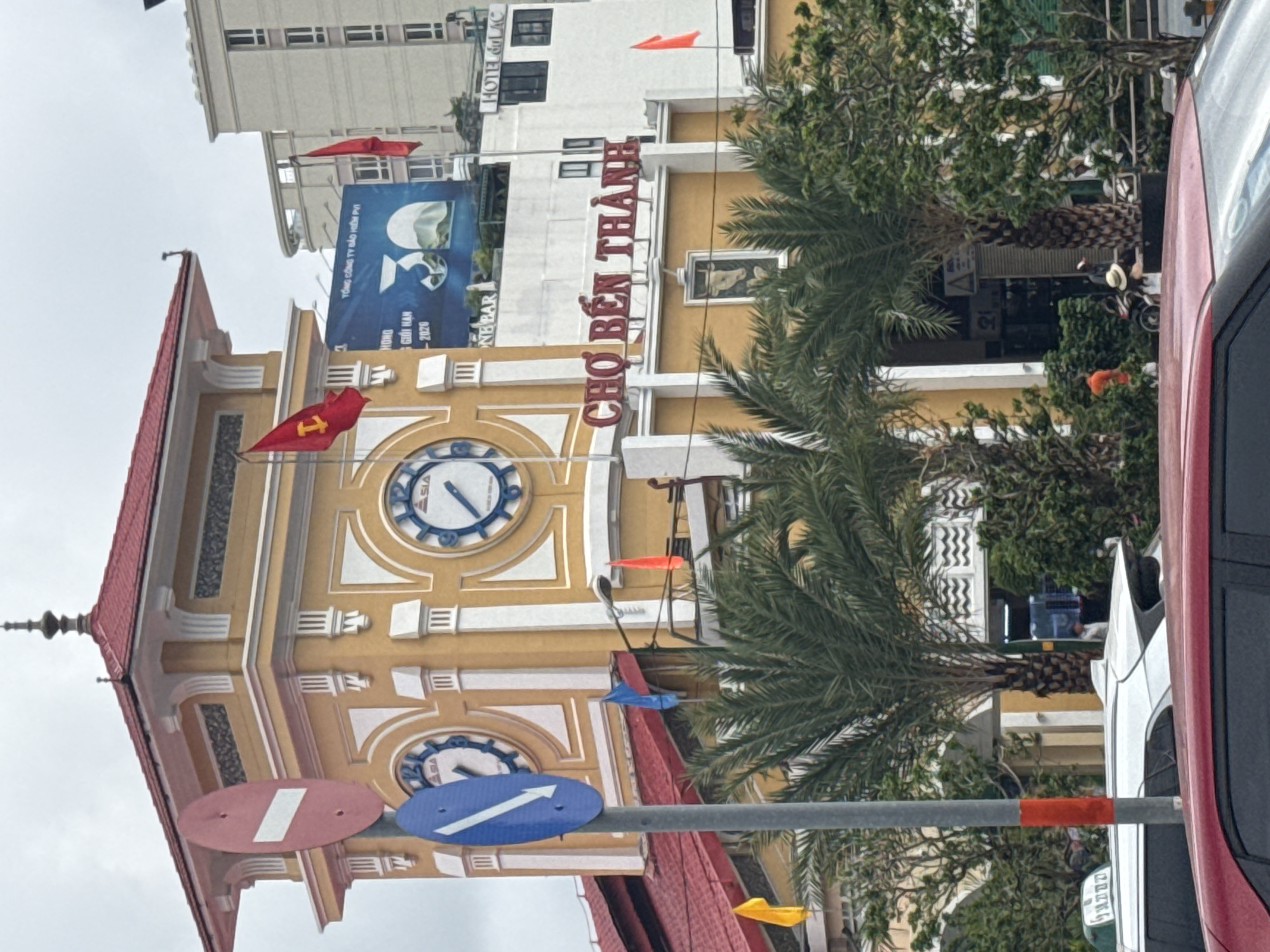 The image shows the Ben Thanh Market in Ho Chi Minh City, Vietnam, a well-known landmark. A large, yellow clock tower with a red roof dominates the scene, featuring two large clocks. In the foreground, traffic signs are visible, including a red "no entry" sign and a blue directional sign. Palm trees and other greenery surround the market. A white car and a red car are partially visible in the lower portion of the image. Several flags, including red and yellow ones, are displayed on poles. A billboard with a blue background and text is mounted on a building behind the clock tower. People are seen in the background, some near entrances to the market and others obscured by trees. The overall environment appears to be a busy urban street.