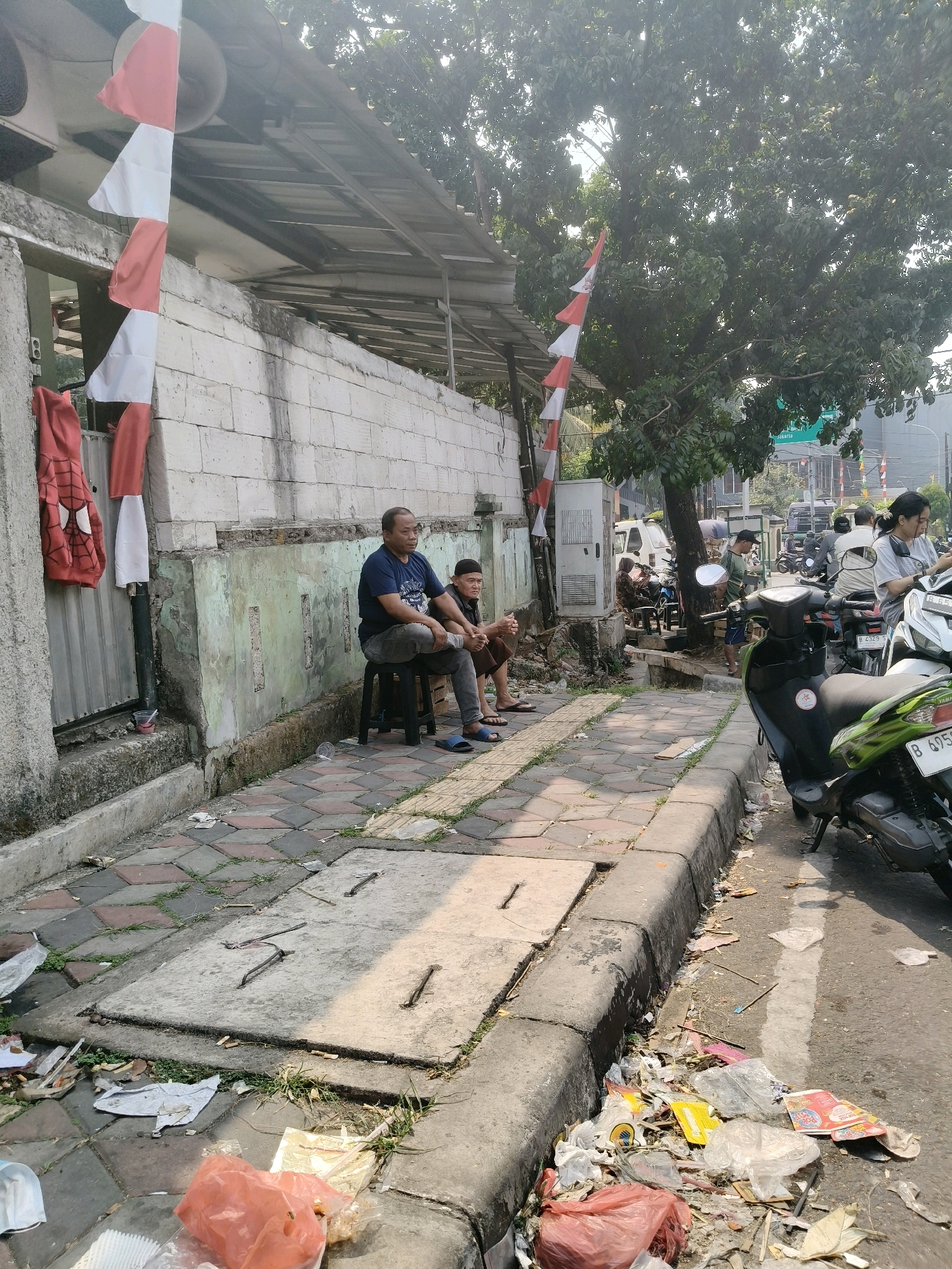 The image depicts a street scene in South Tangerang, Indonesia, with two men sitting on stools on a sidewalk. The man on the left wears a dark blue t-shirt and grey pants, while the man on the right wears a black cap and a dark shirt with brown shorts. Both are facing towards the right of the frame. Behind them, a brick wall is visible, with red and white Indonesian flags hanging vertically. A red jacket with a spider-man logo is draped on a metal pole to the left of the men.
The sidewalk is paved with interlocking bricks and is lined with a concrete curb. The area is littered with trash, including plastic bags, paper, and other debris. Beyond the curb, a street is visible with several motorcycles parked and people on them. A large tree with dense foliage provides shade over part of the scene. In the background, traffic and buildings suggest an urban environment. The lighting indicates it is daytime, with sunlight casting shadows.