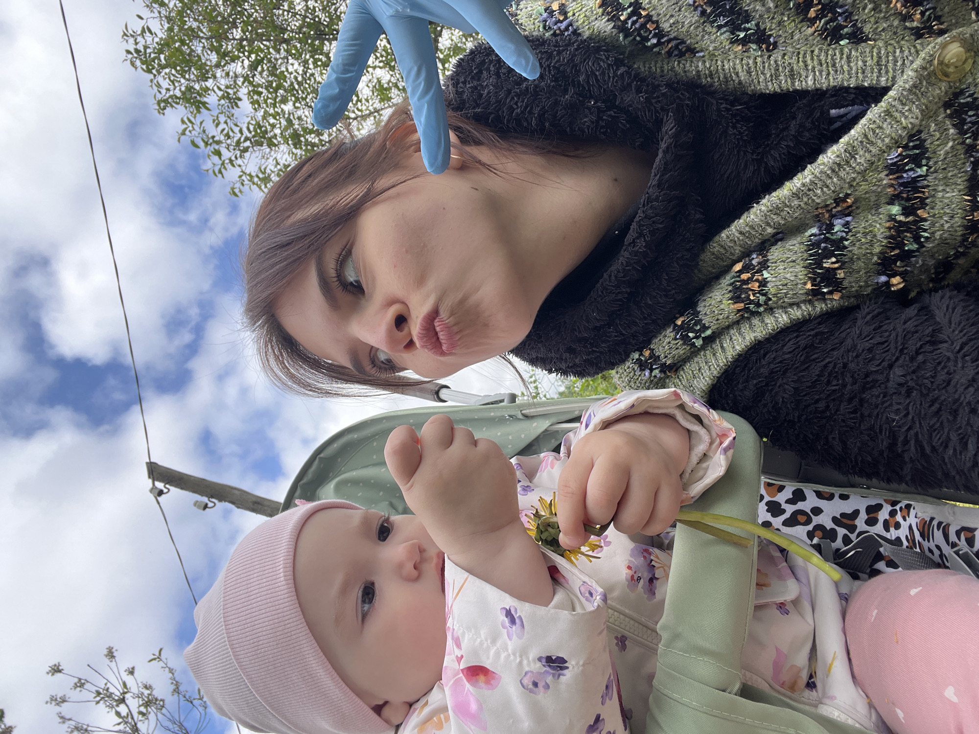 A woman and a baby are in a baby carriage. The woman is wearing a black and green coat and has one hand in a blue glove. The baby is wearing a pink hat and is sitting in a baby carriage with a floral outfit and is holding a dandelion. The image was taken outside, and the sky is visible in the background. The image was taken in Mshinskaya, Russia.