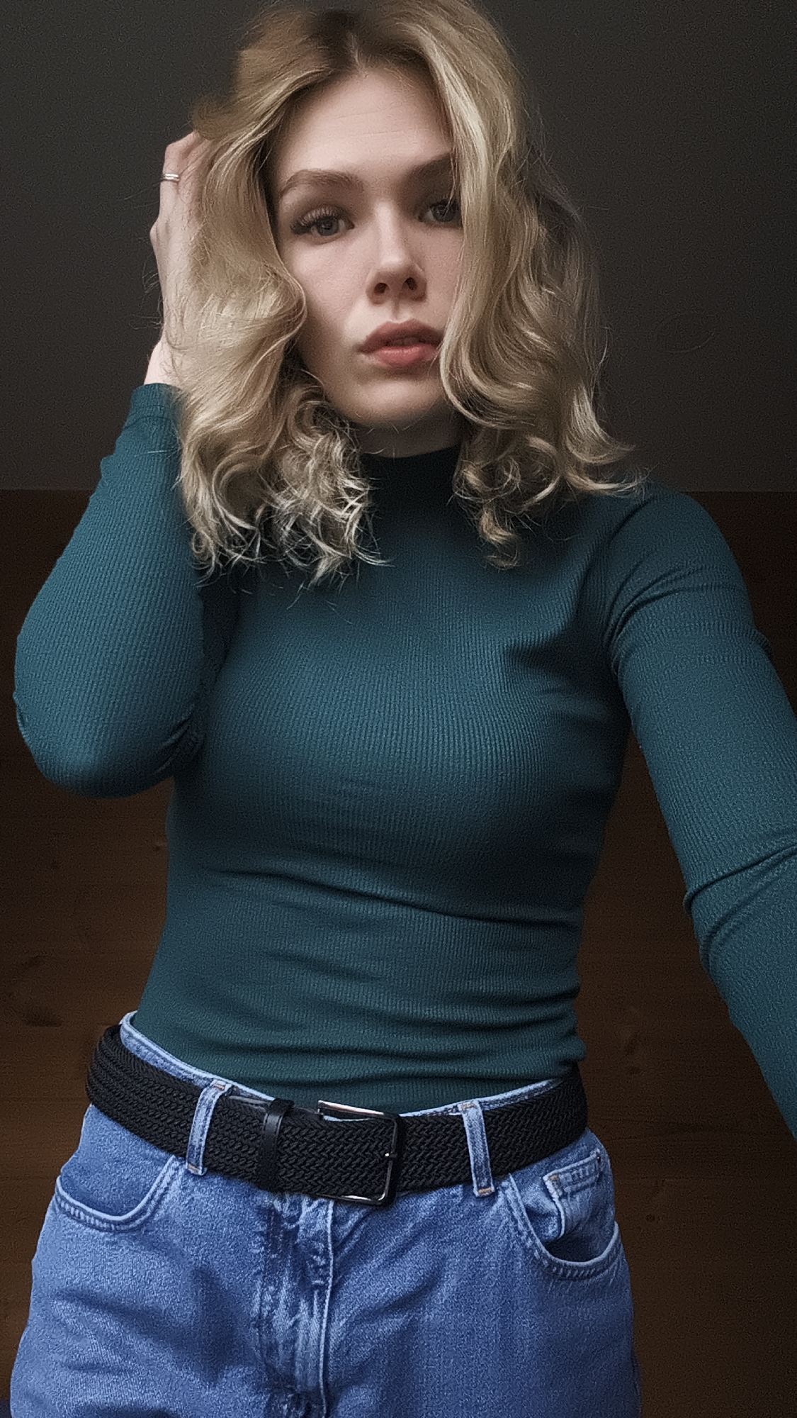 A young woman is posing indoors. She has blonde curly hair and is wearing a green ribbed long-sleeved top, blue jeans, and a black belt. She is looking at the camera and has her hand up near her hair. The background is a dark wall above a light wooden surface. There is no evidence of the location being Русский Вожой, Russia.