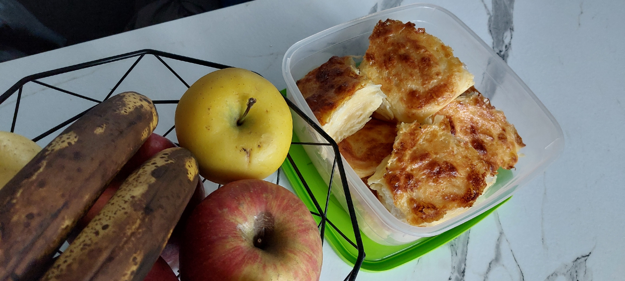 The image captures a still life arrangement of food items on a white, marbled surface, likely a kitchen countertop, in Yagodovo, Bulgaria.
On the left, a black, geometric wire fruit basket contains various produce. Two prominent bananas, showing signs of ripeness with dark brown spots, rest within the basket. Alongside them is a bright yellow apple with its stem, and a red apple with faint stripes, also with its stem intact. A light-colored potato is partially visible behind the bananas.
To the right, a clear plastic food container with a vibrant green base holds several pieces of a baked good. These pieces are golden-brown on top, suggesting a crispy texture, and one broken piece reveals multiple flaky layers of pastry, characteristic of a savory pie such as banitsa or burek, possibly with a cheese filling given the color.
The scene depicts an ordinary indoor setting, possibly a kitchen or dining area, with items typically found in a home. No people are present, and no specific activity is taking place. The lighting is even, indicating an interior shot during the day or under artificial light. There is no visible text in the image.