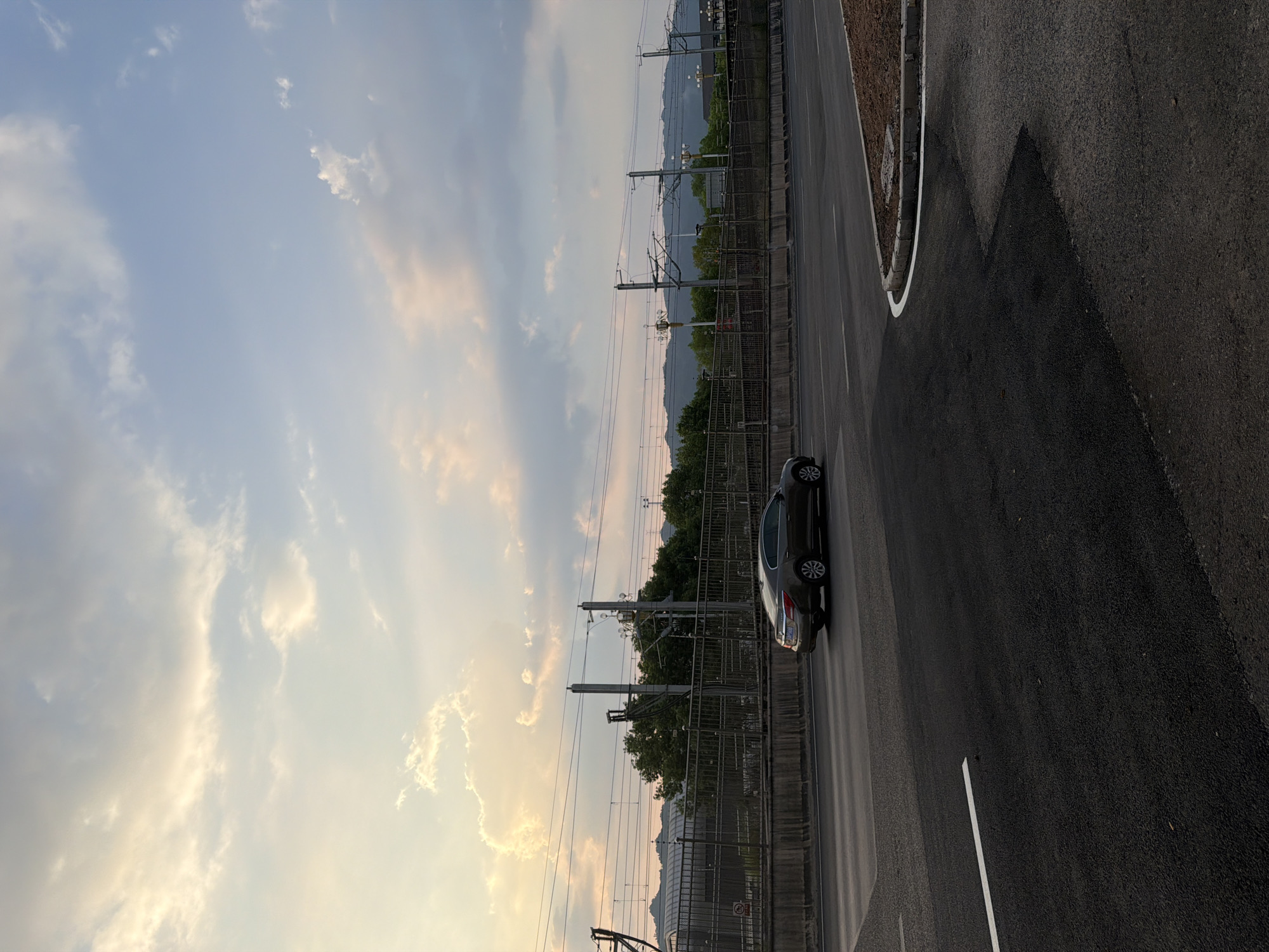 This image captures a serene sunset scene on a highway in Jiangjin, China, likely during the summer months given the lush greenery. The sky is a spectacle of soft hues, with the sun's warm golden light illuminating scattered, wispy clouds, creating a picturesque backdrop against the clear blue sky. In the middle ground, a dark sedan is the sole vehicle visible on the highway, its sleek form captured in motion. Adjacent to the highway, a series of power lines stretch horizontally across the frame, supported by tall pylons, hinting at the presence of infrastructure for the nearby urban area or railway. Beyond the power lines, a dense line of trees forms a verdant barrier, obscuring the view of any structures behind them, with distant mountains forming a hazy silhouette against the horizon. The foreground of the image is dominated by the asphalt of the highway, with a crisp white line marking the lane. To the right, a curved section of the road reveals a newly constructed concrete curb, partially filled with earth, suggesting ongoing roadwork or landscaping. The overall impression is one of tranquil movement and the natural beauty of a rural-meets-infrastructure setting.