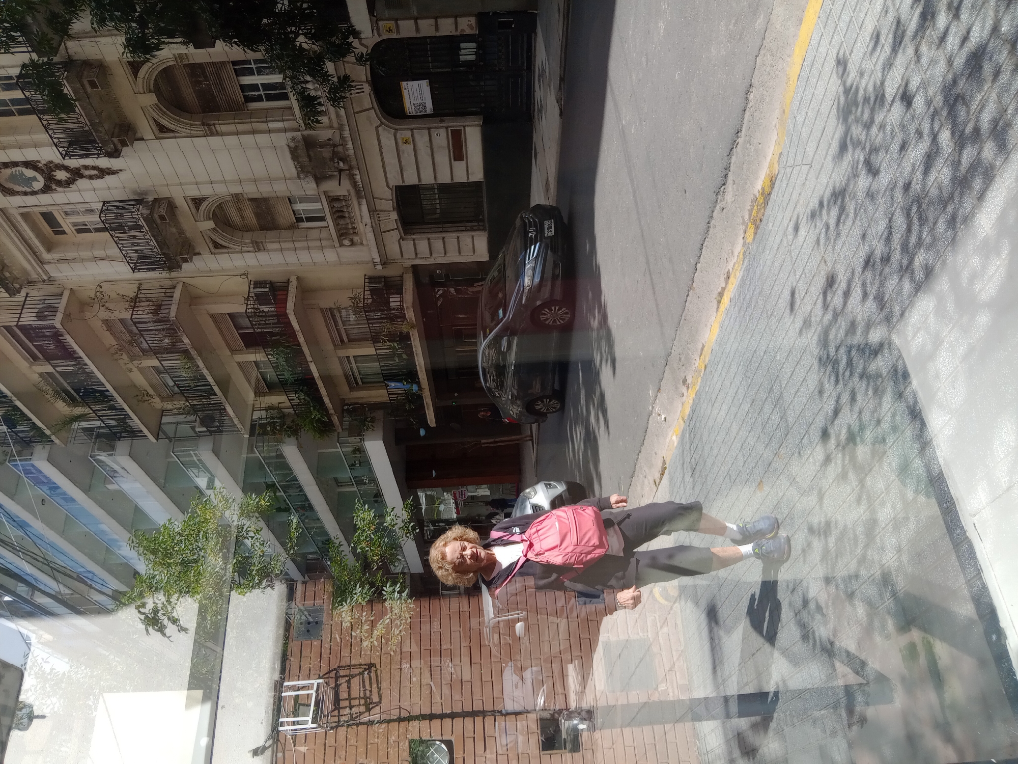 The image shows a woman standing on a sidewalk in Buenos Aires, Argentina, reflected in a window. The street has a parked car and buildings of varying architectural styles, including some with balconies. The sidewalk is paved with a diamond pattern and has a yellow curb. The woman wears dark clothing, sneakers, and a pink backpack. The reflection of the buildings and the sidewalk creates a distorted, layered effect.