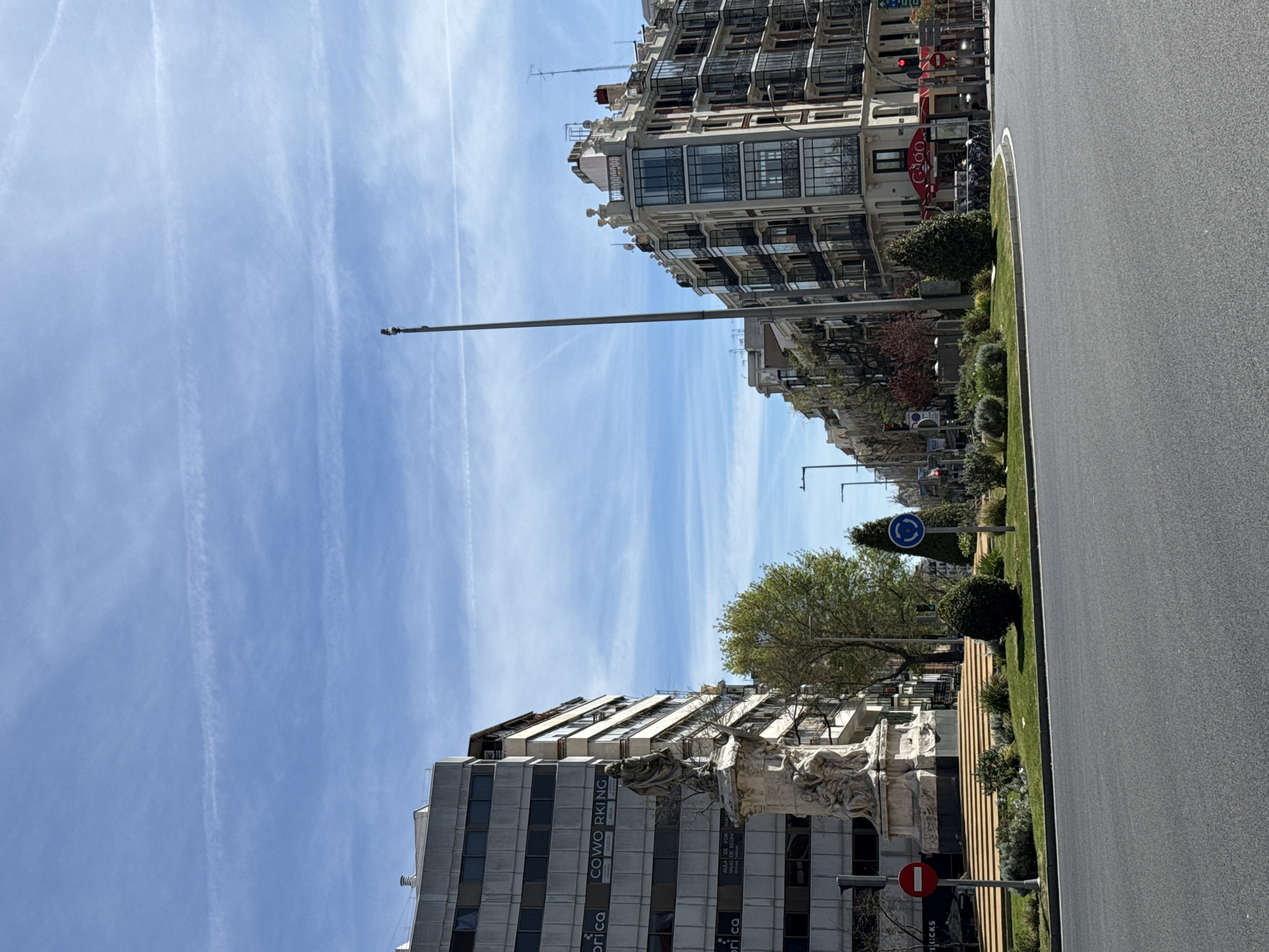 The image depicts a street scene in Madrid, Spain. It is oriented with the horizon running vertically. A wide, gray asphalt road occupies the right side. Greenery, including grass, bushes, and trees, lines the road. Buildings flank both sides of the road, ranging in style from older, ornate architecture to more modern structures. The sky above is blue with white contrails. There are various signs along the road, including a traffic sign indicating a roundabout and a red "no entry" sign. There are people, but they are too small to be identified. One building on the left side has "COWORKING" written on it.