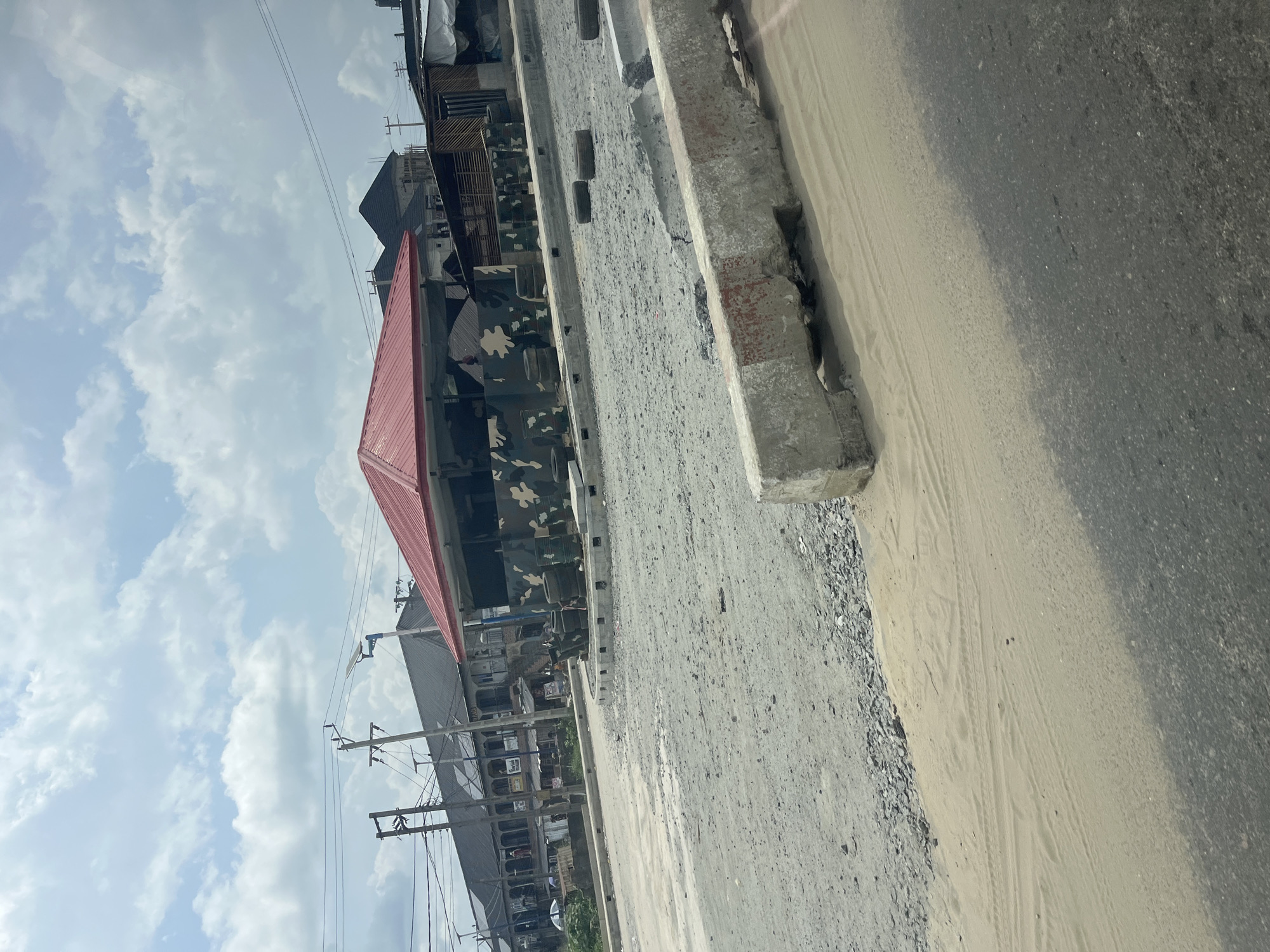 The image presents an outdoor street scene in what appears to be a semi-urban or developing area, consistent with the location context of Asubi, Nigeria. The weather is fair, with a bright sky featuring scattered white cumulus clouds, indicating daytime.
In the foreground, a paved road surface occupies the right side of the frame, partially covered by a layer of light-colored sand and dirt. A sturdy concrete curb or barrier, showing signs of wear and tear, runs parallel to the road on the left. Beyond the curb, there's a rough strip of ground mixed with gravel and dirt.
The mid-ground is dominated by a distinctive building featuring a prominent red sloped roof and walls adorned with a green, brown, black, and white camouflage pattern. Several dark, possibly cylindrical objects (resembling tires or drums) are positioned near this building. Further into the background, other buildings of varying sizes and designs can be seen, suggesting a mix of residential and perhaps commercial structures.
Numerous utility poles with a dense network of overhead electrical wires are visible on the left side, extending into the sky. No people are discernibly present in the scene, and there is no legible text visible on any signs or structures. The image captures a static moment of this street environment, with no particular activity or event taking place.
