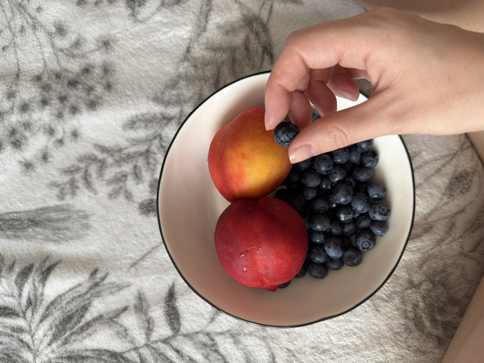 A white bowl filled with blueberries and two nectarines rests on a white blanket with a grey floral pattern. A hand is shown holding a single blueberry. The scene is indoors, on a bed or sofa. There is no information available to determine the location in Kyiv, Ukraine.