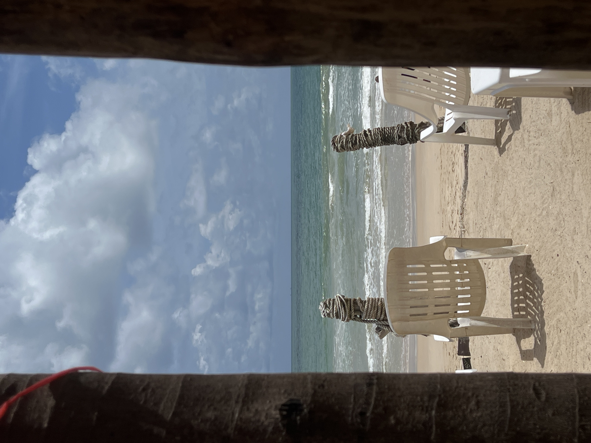 A view of a beach through an opening, likely a window or doorway. The sky is blue with scattered white clouds. The ocean is visible with small waves breaking along the shoreline. Two white plastic chairs are on the sandy beach. A dark, rough-hewn frame surrounds the view, with a bit of red string visible on the lower left side. The location is Unknown, Unknown.