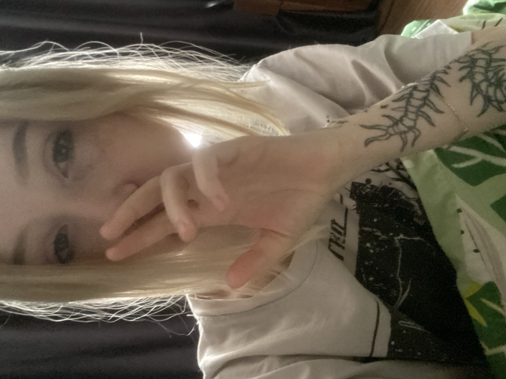 A single individual, appearing to be a young adult female with blonde, shoulder-length hair and light-colored eyes, is depicted in a close-up indoor shot. She is wearing a light-colored short-sleeved t-shirt with a dark, abstract graphic print on the chest. Her left hand is raised to partially obscure the lower half of her face, with fingers slightly parted. The visible portion of her right forearm, positioned in the lower right, displays a dark-ink tattoo resembling a segmented, insect-like or centipede-like creature, along with a thin, light-colored bracelet. The background is dark, featuring a dark curtain or fabric on the left and a darker, possibly wooden furniture item on the right. The lower foreground shows a green and white patterned blanket or fabric. The scene is front-lit, with a bright light source originating from the right, creating highlights on the hair and skin. There are no identifiable landmarks or environmental cues to determine the location within the city of Izhevsk, Russia, or any specific geographic region.