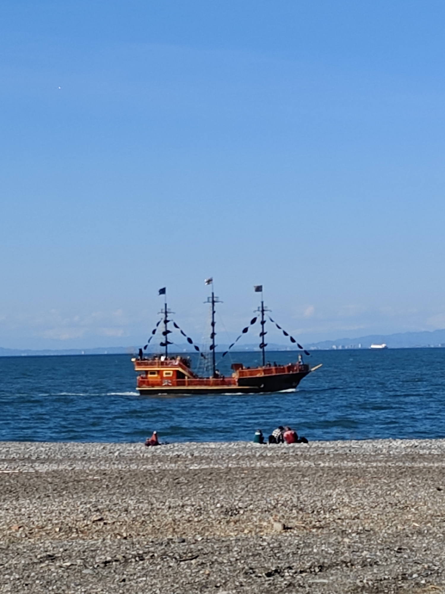 The image depicts a pirate ship sailing on the ocean, with a rocky beach in the foreground. People are gathered on the beach. The sky is clear and blue. A city skyline is visible in the distance. The location appears to be in the city of Unknown, Unknown.