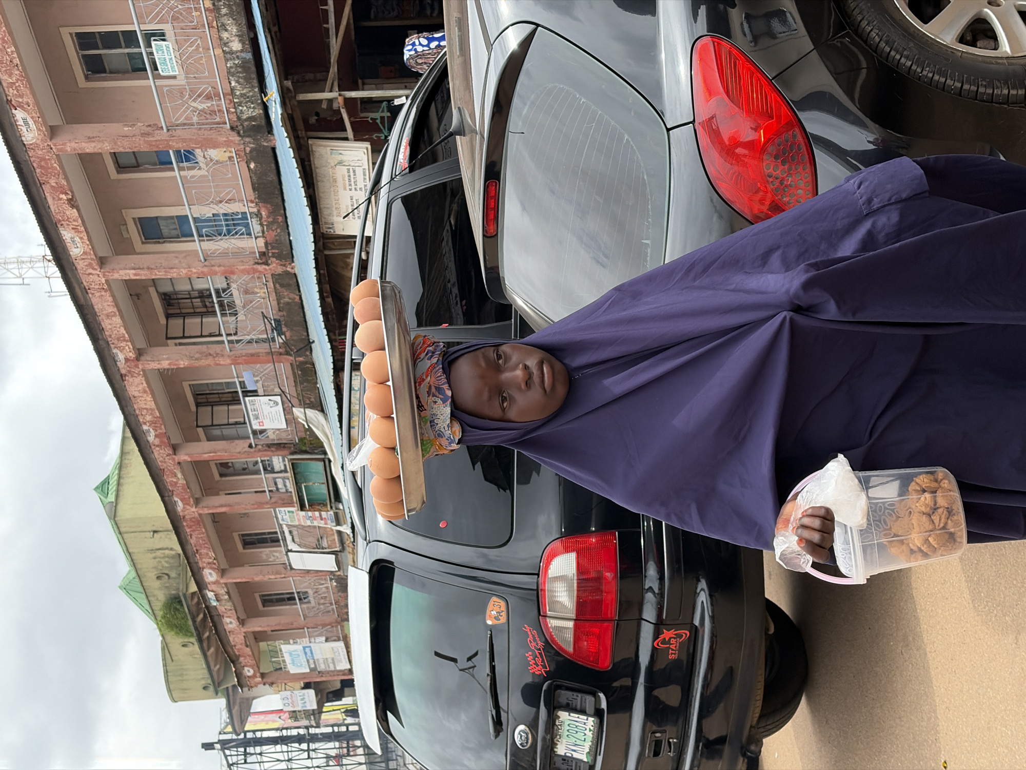 The image depicts a young girl in Jos, Nigeria, carrying a tray of eggs on her head and a bucket of snacks in her hand. She is standing on a street in front of a row of buildings, with cars parked on either side. The buildings appear to be shops or residential units, with balconies and advertisements visible on their facades. The street is paved and there are utility poles and wires in the background. The girl is wearing a purple hijab and a purple garment that covers her body. The eggs are arranged in a single layer on a metal tray. The bucket contains small, round, brown snacks. The overall scene suggests a bustling marketplace or a busy street in a Nigerian city.