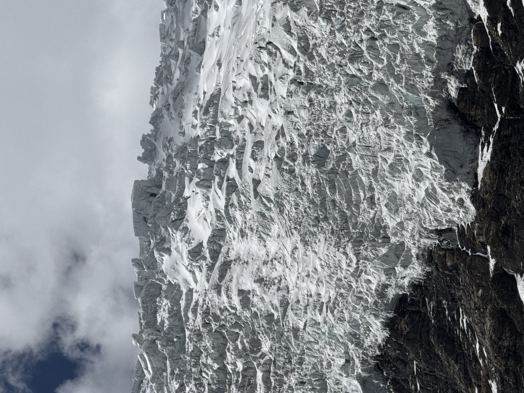 The image captures a close-up view of a steep, snow-covered mountain face in Gyantse County, China. The mountain consists of light grey, heavily textured glacial ice and rock. The ice appears brittle and cracked, with numerous crevices and ridges. To the right, a dark, rugged rock face is visible, contrasting with the bright snow and ice. The sky is overcast with light grey clouds in the left portion of the frame, with a patch of dark blue breaking through.
