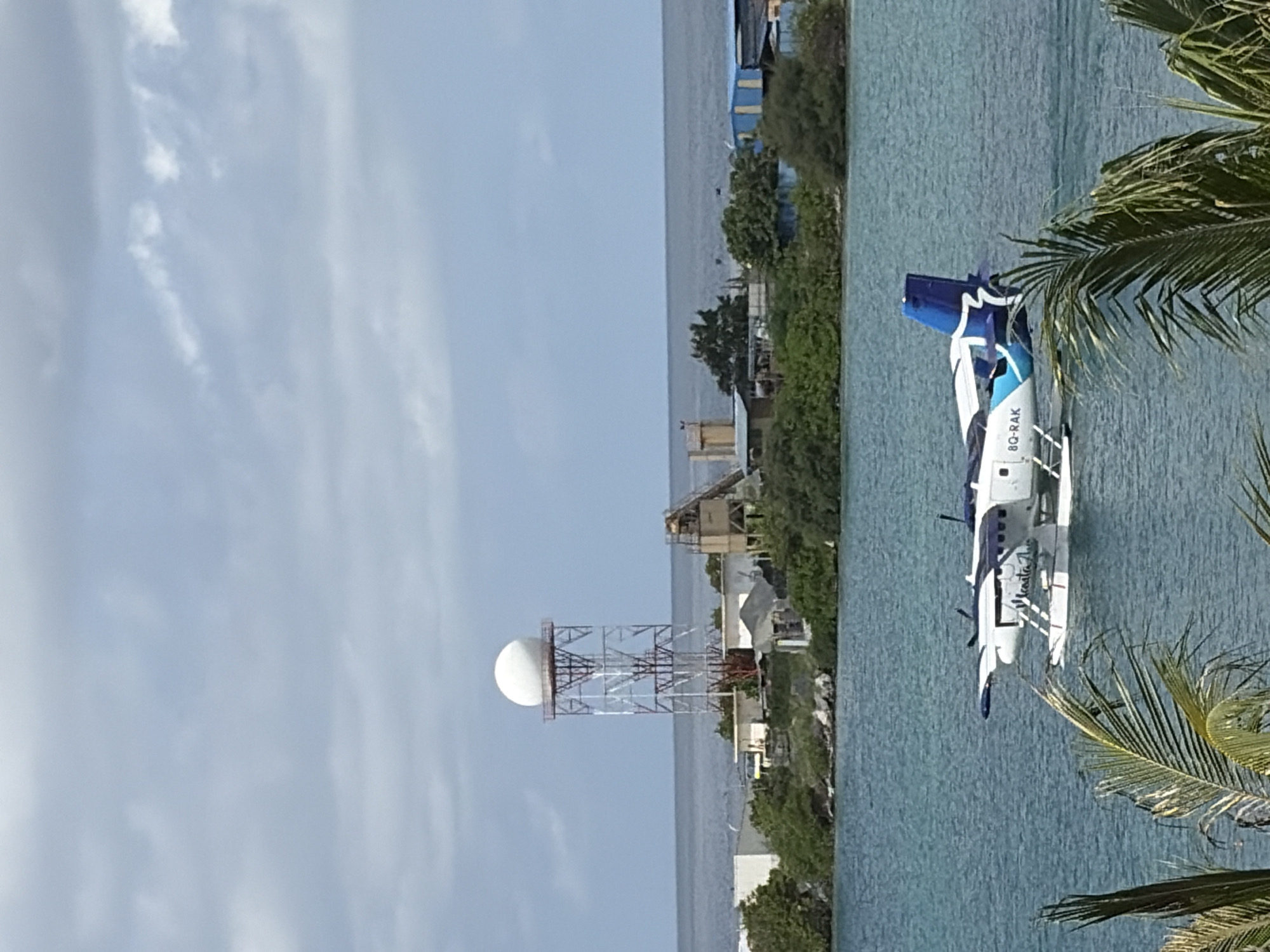 A seaplane with the registration "8Q-RAK" is on the water in front of a coastal area of Malé, Maldives. The seaplane is white with blue accents and has two propellers on top of its wings. In the background, there is a radar tower on a hill, surrounded by buildings and lush green vegetation. The sea is calm and blue, with a cloudy sky overhead. There are palm fronds in the foreground.