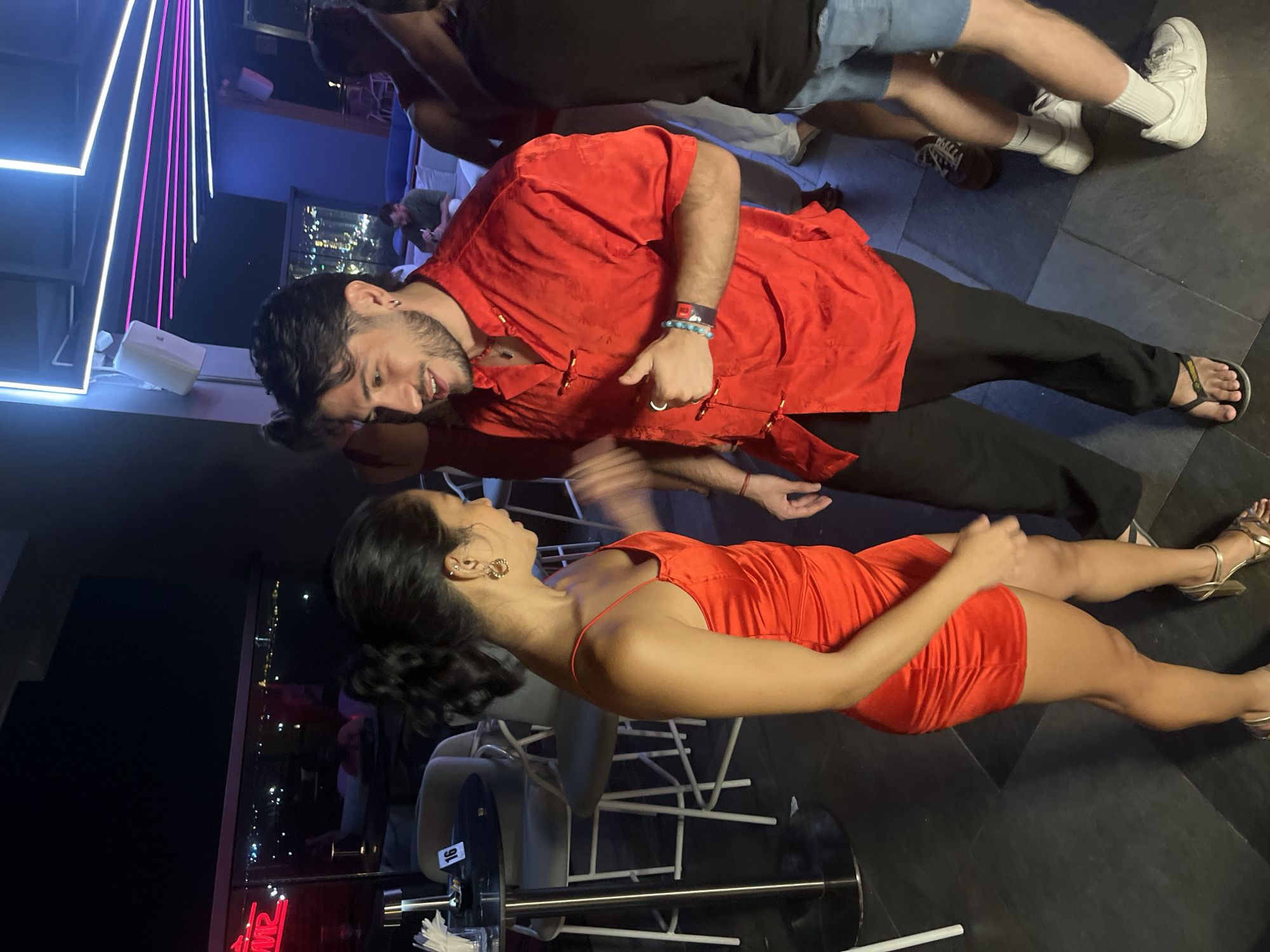 The image depicts a nighttime scene on a rooftop bar in Da Nang, Vietnam, with the city lights visible in the background. In the foreground, a couple is interacting closely. The woman is wearing a short, strapless red dress and gold heels, with her hair pulled back in a ponytail. The man is dressed in a red, oriental-style shirt with frog closures and black pants, accessorized with a beaded bracelet. They appear to be engaged in conversation or dancing, with a celebratory atmosphere suggested by the neon lighting and the presence of other patrons in the background. The bar area features high-top tables and chairs, typical of a modern establishment.