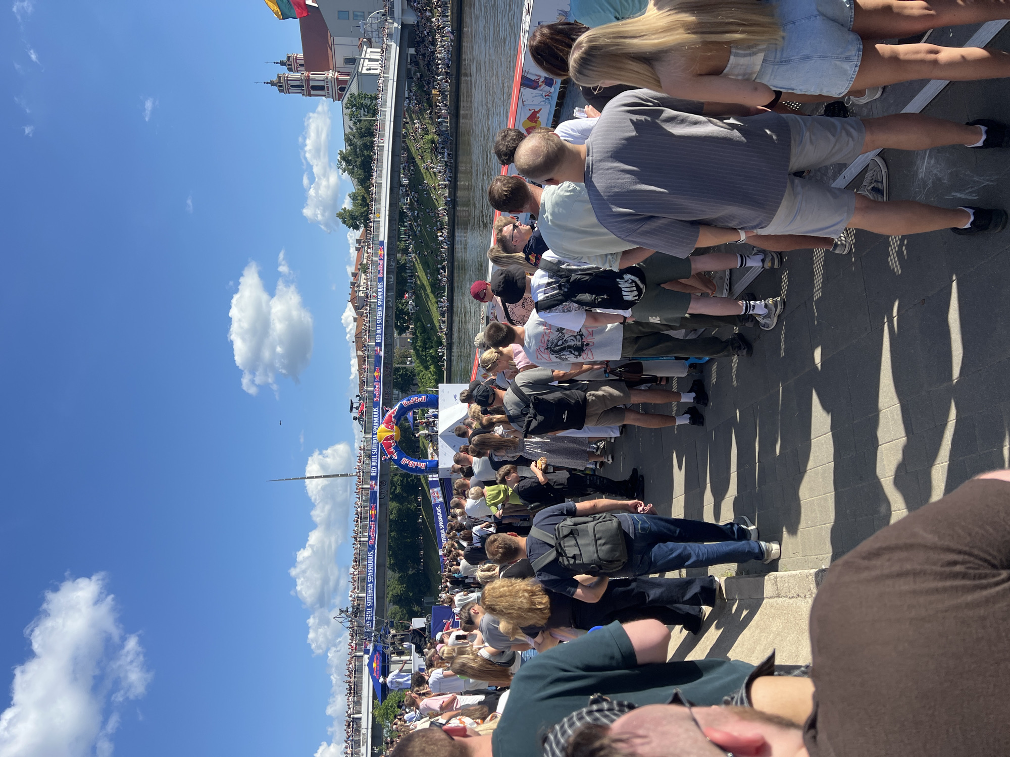 The image depicts a large gathering of people on a sunny day, likely in Vilnius, Lithuania, given the architectural style of the church in the background and the presence of a Red Bull sponsored event. The scene is set along a waterfront, with a river visible on the right side of the frame. A bridge stretches across the river in the middle ground, crowded with spectators, and a Red Bull archway is visible on the bridge, indicating a sporting or entertainment event.
In the foreground, a long queue of people is waiting, many with their backs to the camera. They are dressed casually in summer attire. Some individuals are wearing backpacks. The ground is paved with interlocking stones, and shadows are cast by the sunlight, creating a patterned effect.
In the middle ground, beyond the queue, there is a stage area with the Red Bull archway as a prominent feature. More people are visible on and around the stage. Further back, the bridge is packed with a dense crowd, and a building with distinctive twin towers, resembling a church or cathedral, stands on the right side of the frame in the background. The sky is blue with scattered white clouds. The overall impression is of a lively outdoor event attended by a significant number of people.