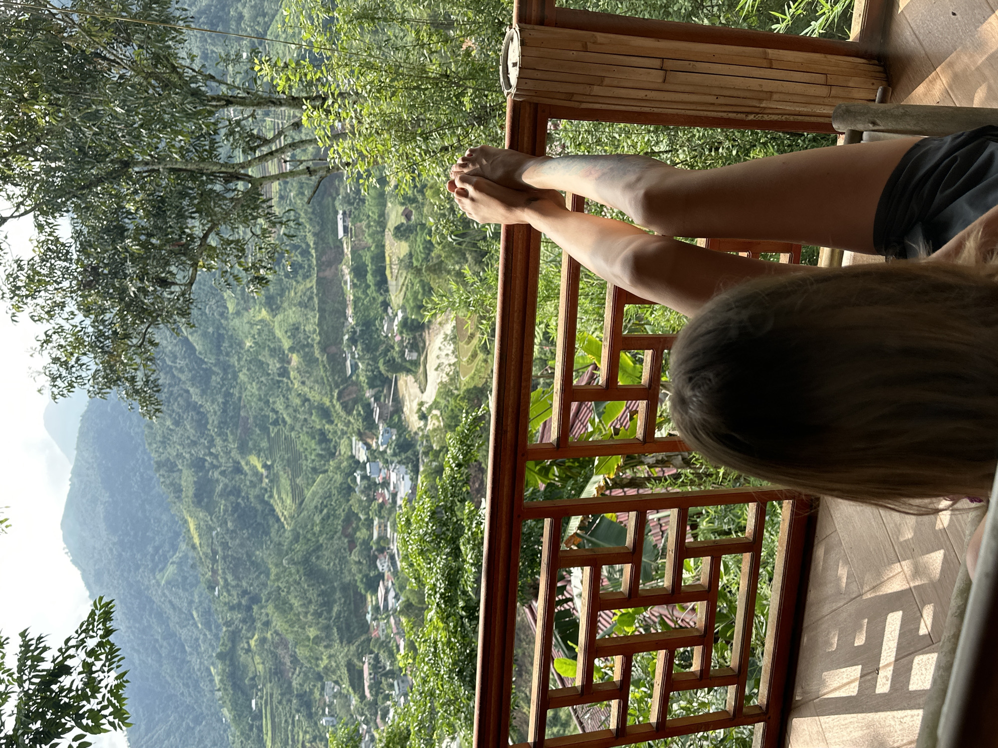 The image captures a serene outdoor scene from a balcony overlooking a vast, verdant mountain landscape in Thông Nguyên Commune, Vietnam. A person, likely female, is featured in the foreground, seen from behind and from the torso down. She is relaxing and facing away from the camera, with her legs, clad in dark shorts, propped up on a wooden balcony railing, suggesting a posture of leisure and comfort. Her long, light brown hair is visible from behind her head.
The balcony itself is constructed with dark, ornate wooden railings featuring intricate geometric patterns, and a lighter-toned wooden floor. Sunlight streams onto the balcony, casting distinct shadows of the railing onto the floor, indicating it is daytime with clear weather, possibly morning or late afternoon. A rolled-up bamboo blind is visible on the upper right side of the balcony, and lush green foliage and trees are abundant in the immediate surroundings.
Beyond the balcony, a breathtaking panorama unfolds. Rolling green mountains covered in dense foliage and terraced fields stretch into the distance under a bright sky. Scattered small buildings and structures, indicative of a village, are nestled in the valley below and along the mountain slopes. The scene evokes a sense of tranquility and appreciation for the natural beauty of the Vietnamese highlands. There is no visible text in the image.