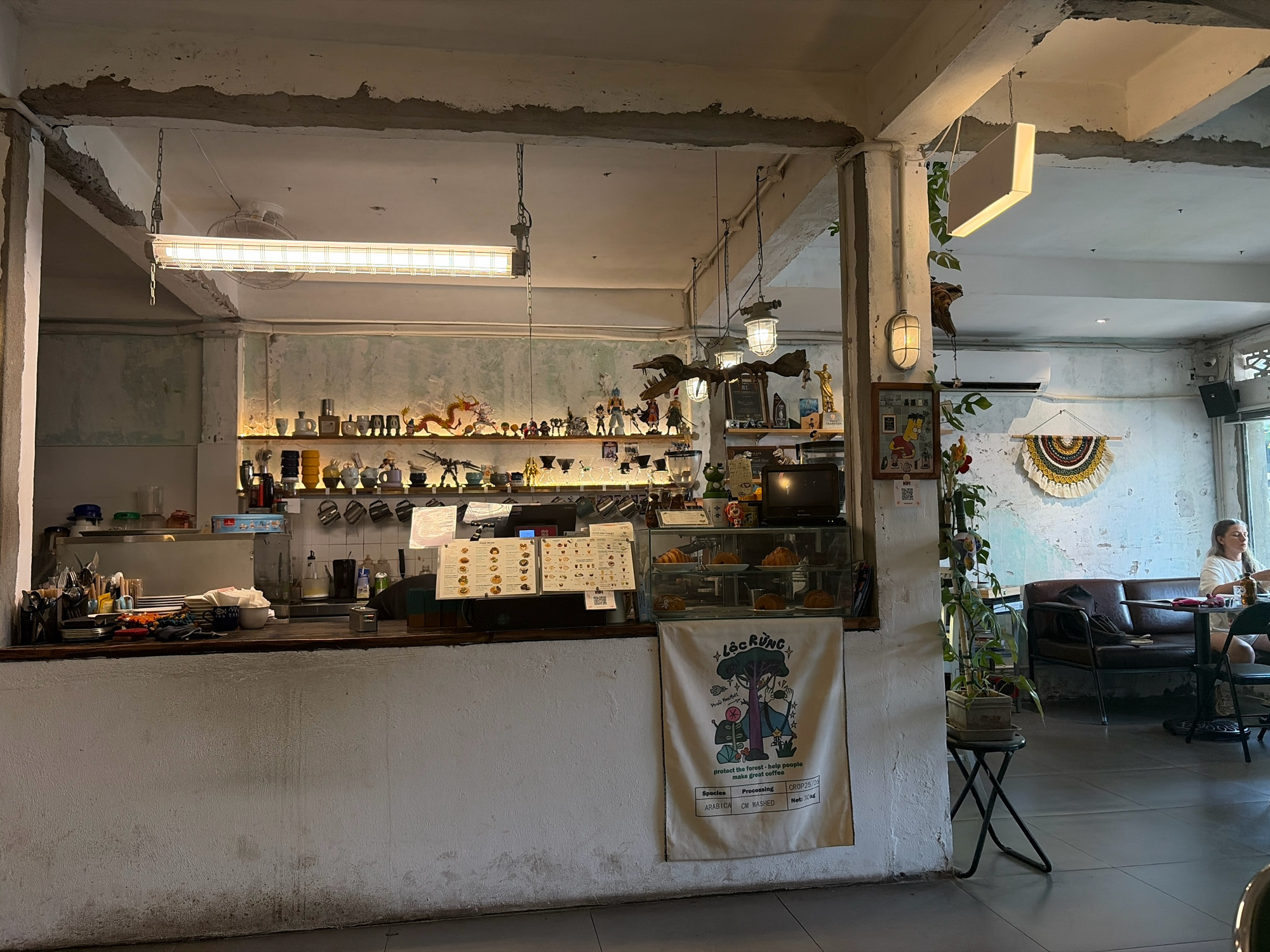 The image shows the interior of a coffee shop in Da Nang, Vietnam. The shop has a rustic and industrial design with exposed concrete elements. The foreground features a counter area with a display of pastries, coffee machines, and various cups and mugs on shelves above. A white banner with colorful illustrations and text is hanging from the counter. In the background, a woman is seated at a table, and a sofa is visible. The overall lighting is dim and ambient, with overhead fluorescent lights and decorative lamps.