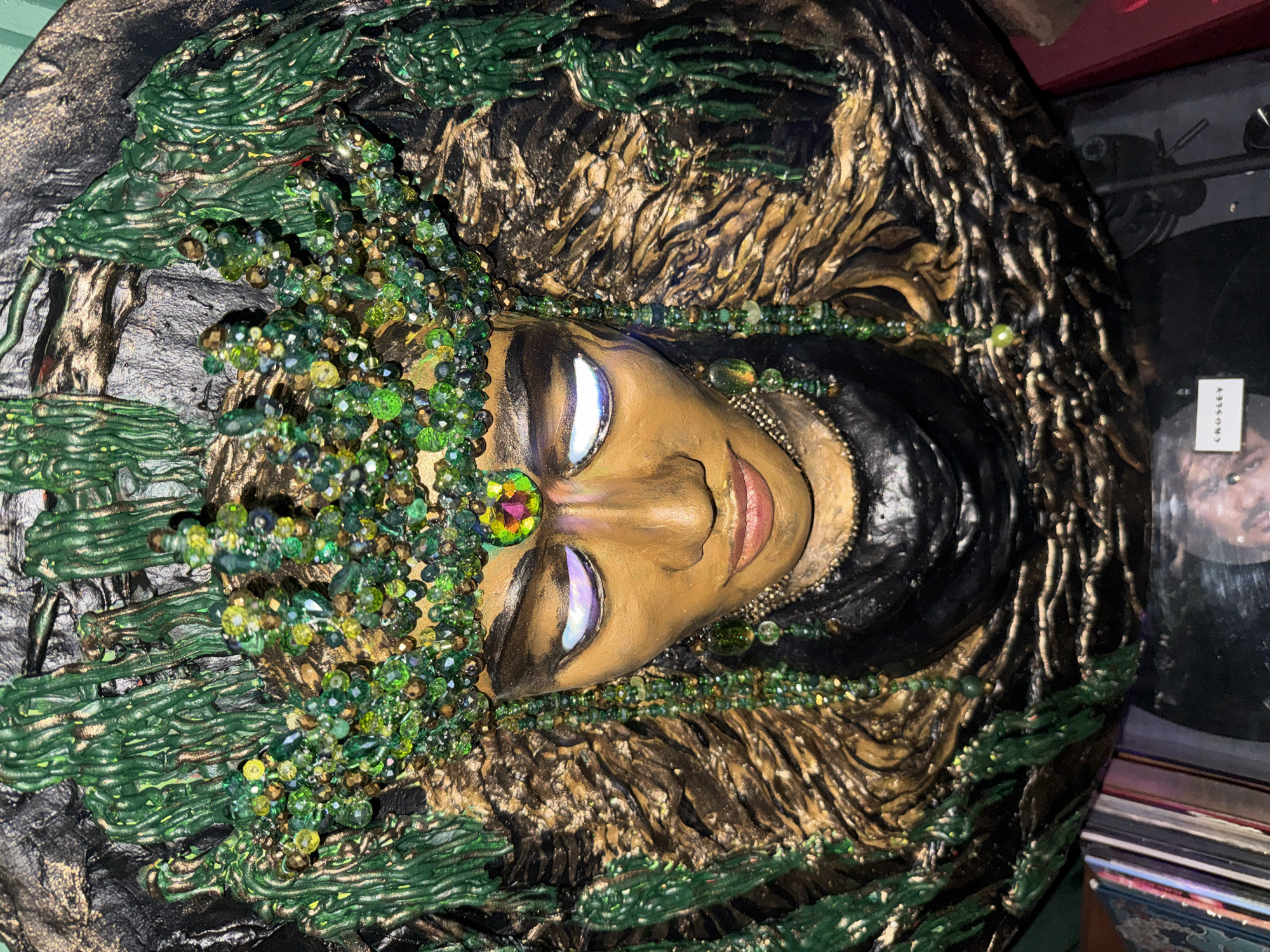 A close-up, low-angle, eye-level shot shows a richly decorated, mask-like artwork of a woman's face. The artwork is predominantly gold and green. The face has a golden complexion with heavily made-up eyes featuring white and purple eyeshadow. A vibrant green and gold jeweled crown adorns her head, with beaded green strands hanging down like hair. The background is textured and dark, with sculpted green elements resembling dripping vines or moss. The artwork appears to be mounted on a circular base. Below the artwork, a stack of records is visible, along with a sliver of a red book or box on the right edge. The overall impression is one of opulence and mystique. The image does not provide any information about the location in Kuta Selatan, Indonesia.