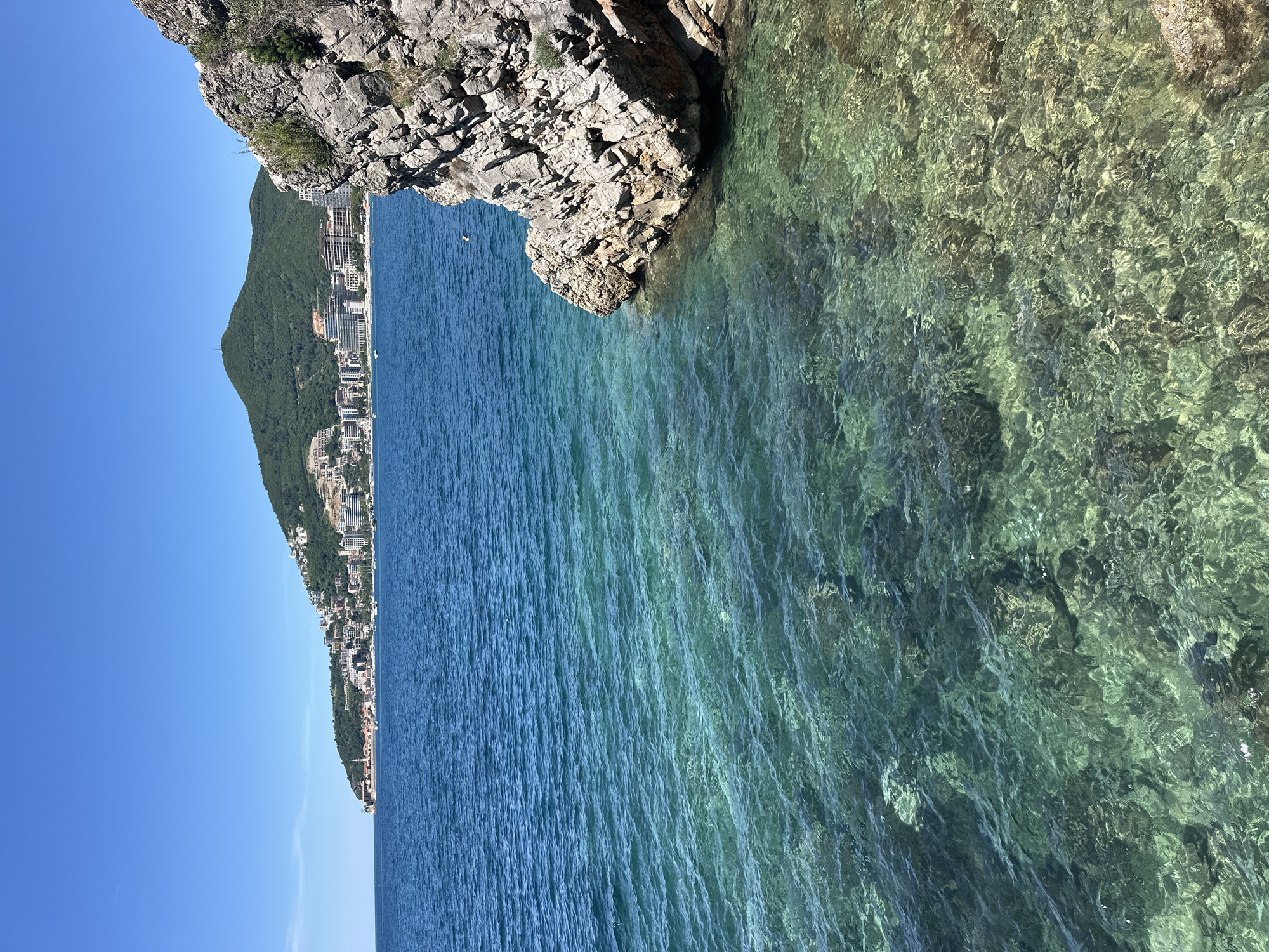 The image depicts a coastal scene in Budva, Montenegro, taken from a high vantage point. The foreground features rocky cliffs on the upper right, transitioning into clear, turquoise water. The clarity of the water allows visibility of the seabed's details. In the distance, the coastline curves, revealing densely built-up areas, likely urban developments and buildings nestled against a backdrop of green hills. The sky is clear and blue. There are no people visible.