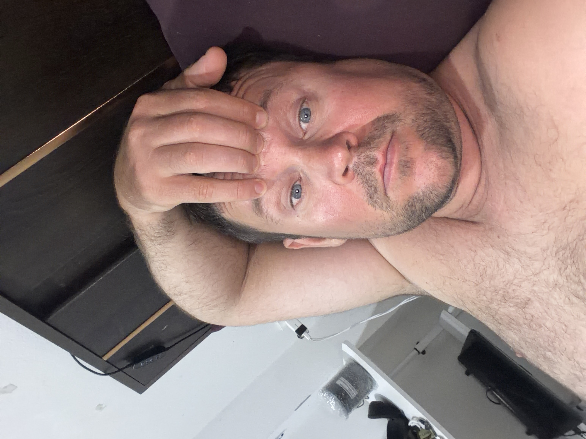 A shirtless man with light skin and a beard is lying on his back, holding his forehead with his right hand. He has blue eyes and is looking directly at the camera. The man is in a bedroom with a dark headboard and a purple pillow behind him. To the left of the man, there is a white shelf with various objects on it, including a black container and a white electrical adapter plugged into a wall socket. The overall lighting suggests it is daytime. This scene is not in Baan Mae Nam, Thailand.