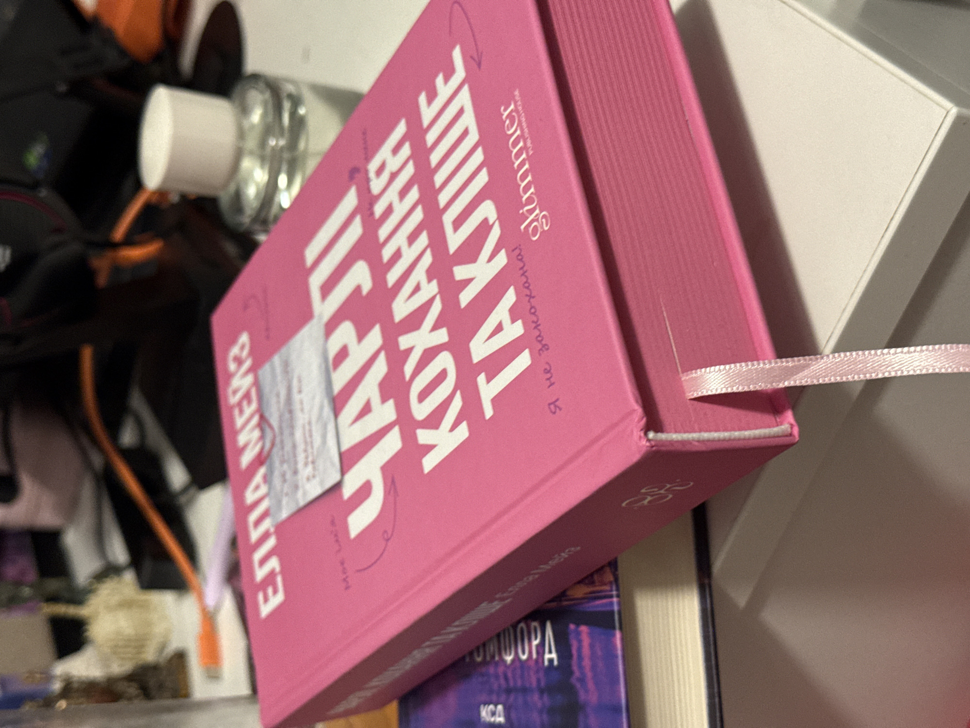 The image shows a close-up, slightly angled view of a bright pink hardcover book with white text. The title in large white Cyrillic letters reads "ЧАРЛІ КОХАННЯ ТА КЛІШЕ". Below this, in smaller text, it says "Я не закохана!". To the right of this text, the word "glimmer" appears in a stylized font, with "PUBLISHING HOUSE" in smaller letters underneath. The book has a pink ribbon bookmark extending from the spine. The book is leaning against another book with a dark purple cover that has some visible text in white Cyrillic. The scene is set on a light-colored surface, possibly a desk or shelf, with the background blurred and showing indistinct objects and lighting, including what appears to be a bottle of liquid with a white cap. The lighting suggests an indoor setting, possibly a room in Łódź, Poland, given the Cyrillic text on the book. There are no people visible in the image.