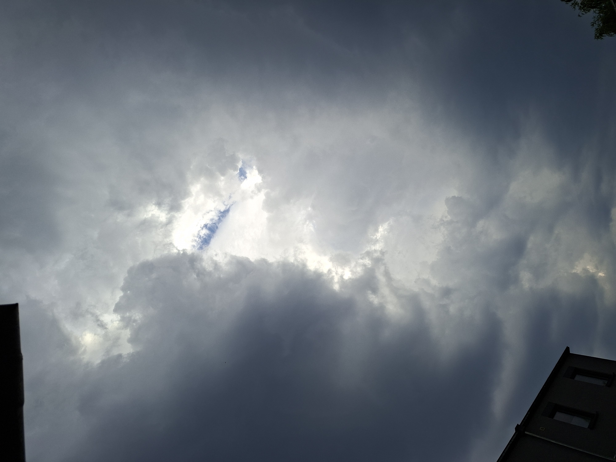 The image shows a dramatic sky with dark, heavy clouds parting to reveal a bright patch of blue sky and sunlight. The clouds are textured and appear to be moving, creating a sense of dynamic weather. In the lower left corner, a portion of a building with two windows is visible, suggesting an urban or suburban setting. The overall impression is one of an impending storm or the aftermath of one. The location is specified as Baden-Baden, Germany.