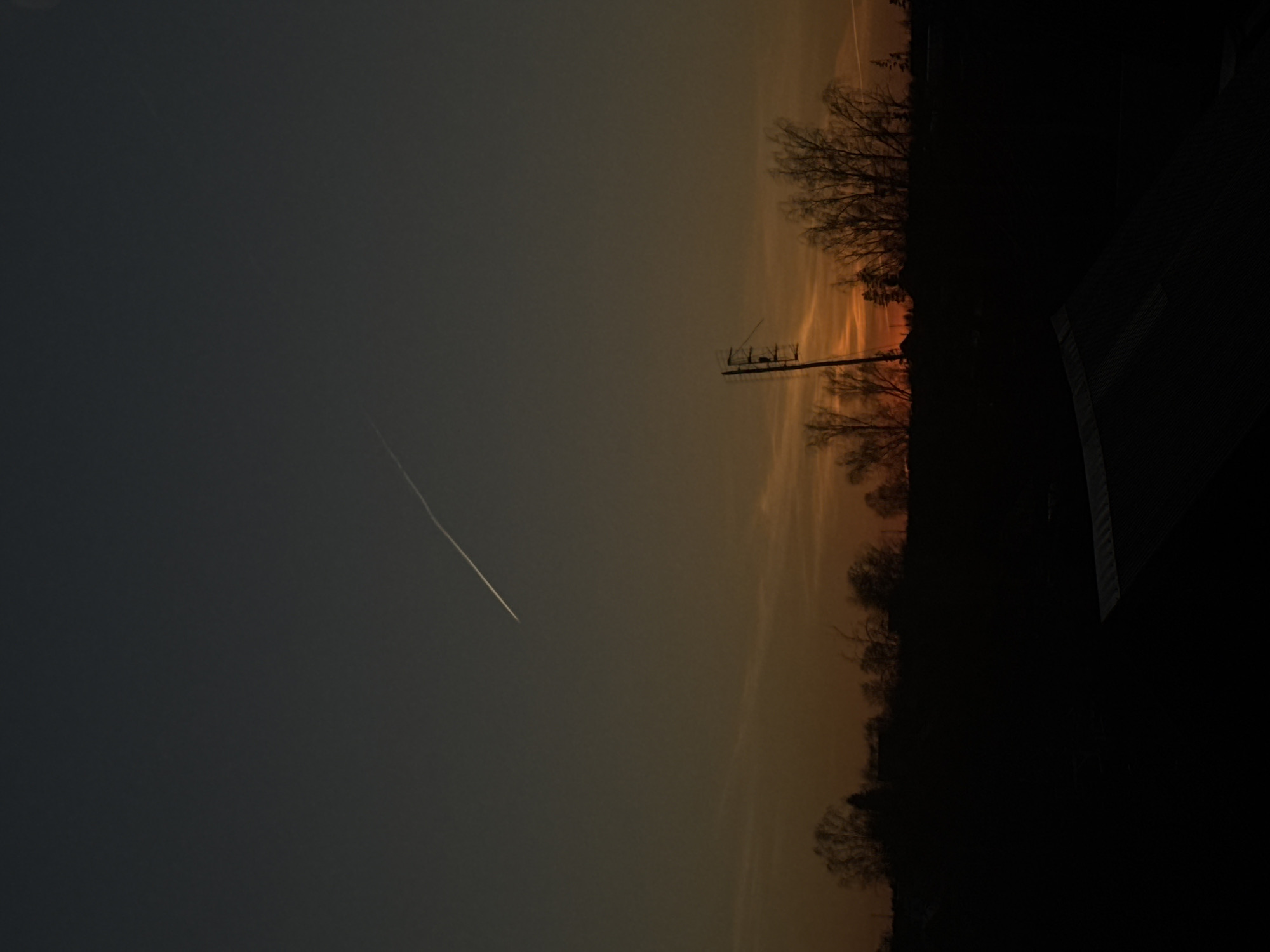 The image shows a sky, transitioning from a darker gray to an orange hue near the horizon. A jet contrail is visible in the darker portion of the sky. Silhouetted trees line the horizon. A utility pole is positioned slightly to the left of the center. A dark, indistinct structure is in the lower right. The image does not appear to be in Фоки, Russia.