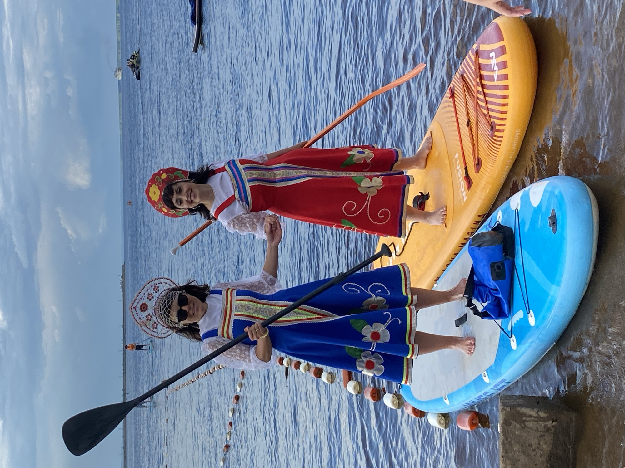 Two women in traditional Russian attire, one in a blue sarafan and the other in a red sarafan, stand on separate paddleboards in the Kama River in Chaikovsky, Russia. Both women wear elaborate headdresses. The woman on the left is holding a paddle, and the woman on the right is also holding a paddle. They are facing the camera and appear to be smiling. In the background, other people are visible on paddleboards and a boat is in the distance. The sky is cloudy.